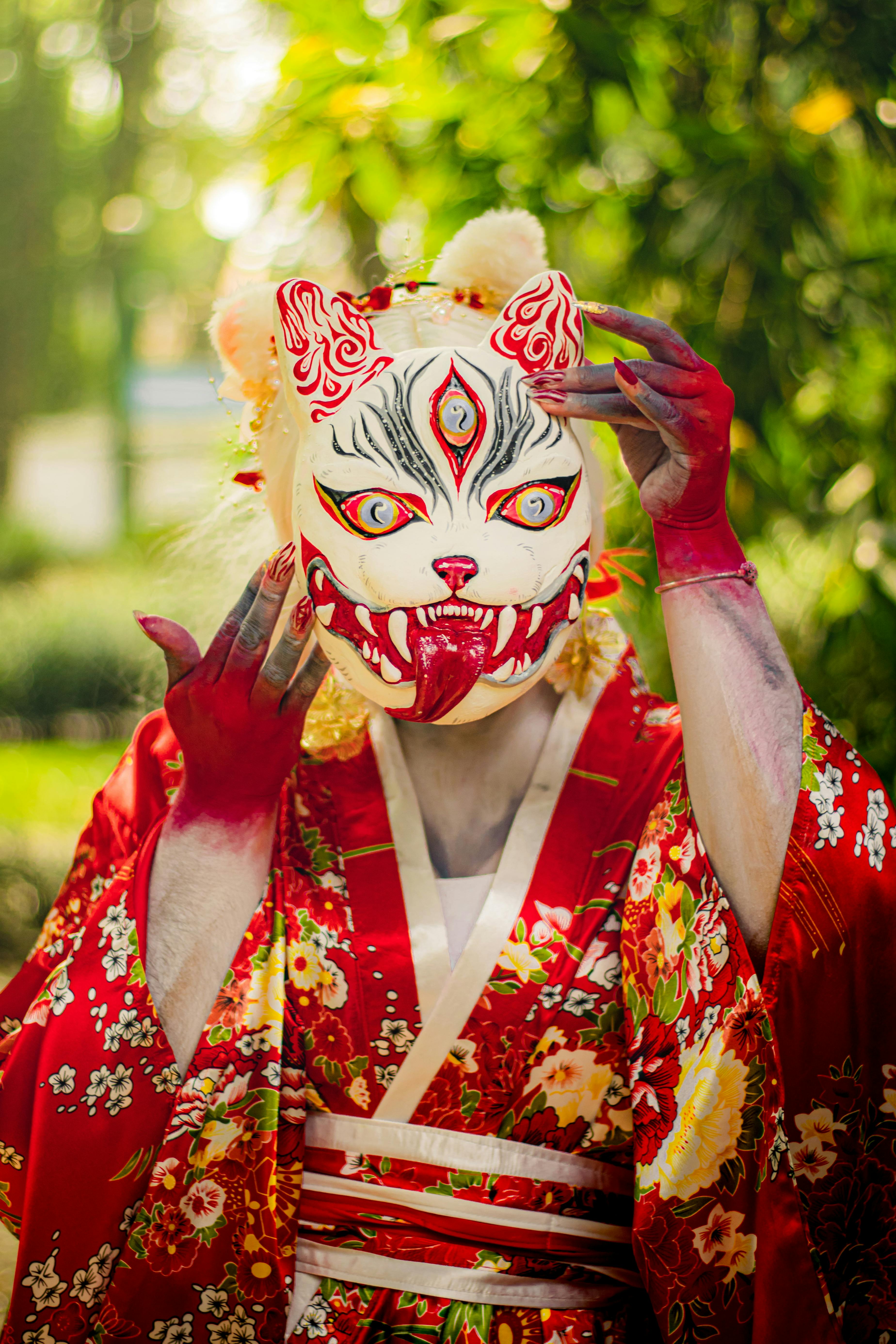 ฟรี บุคคลในชุดกิโมโนสีสันสดใส สวมหน้ากากจิ้งจอกที่ออกแบบอย่างมีศิลปะ ในสวนสีเขียวชอุ่ม คลังภาพถ่าย