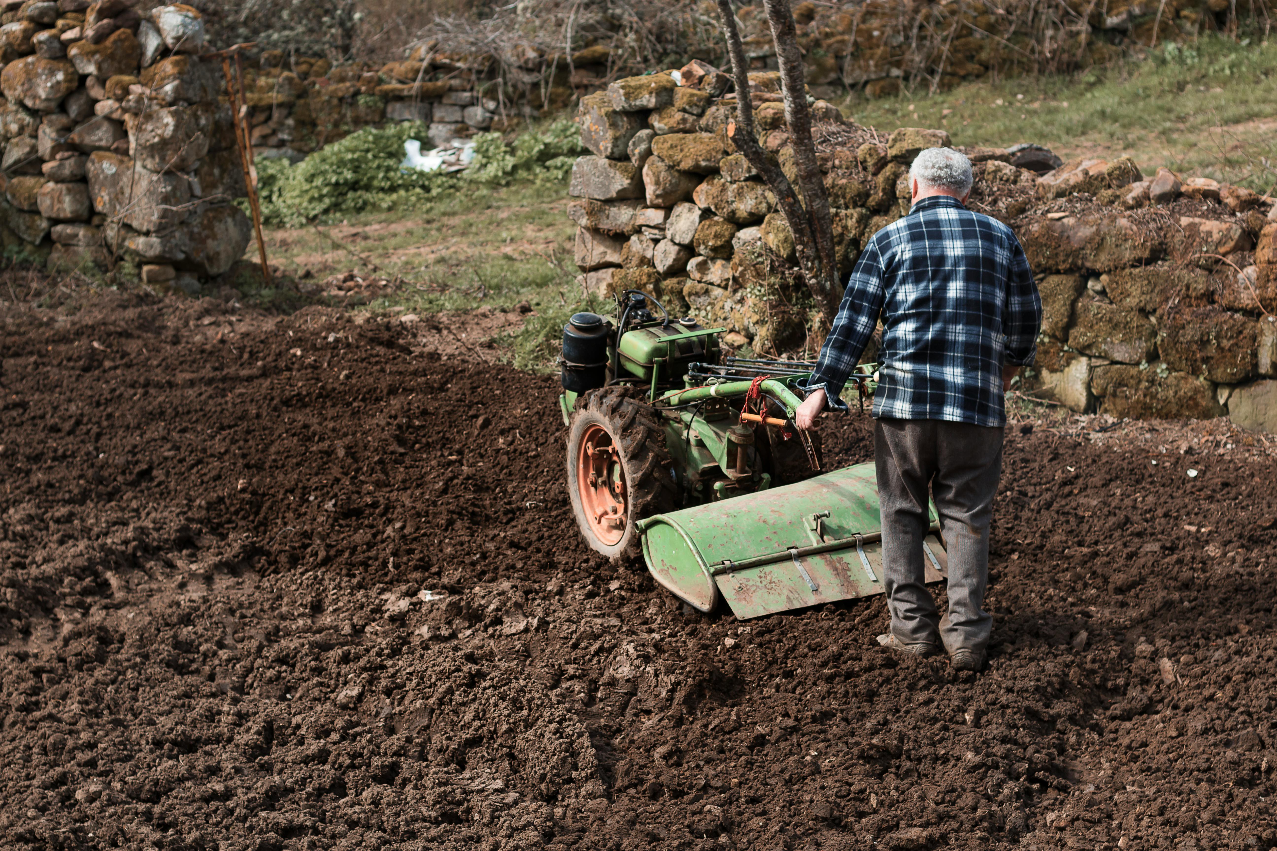 Elderly man using a green tiller in a rustic field with stone walls, embodying traditional farming.
