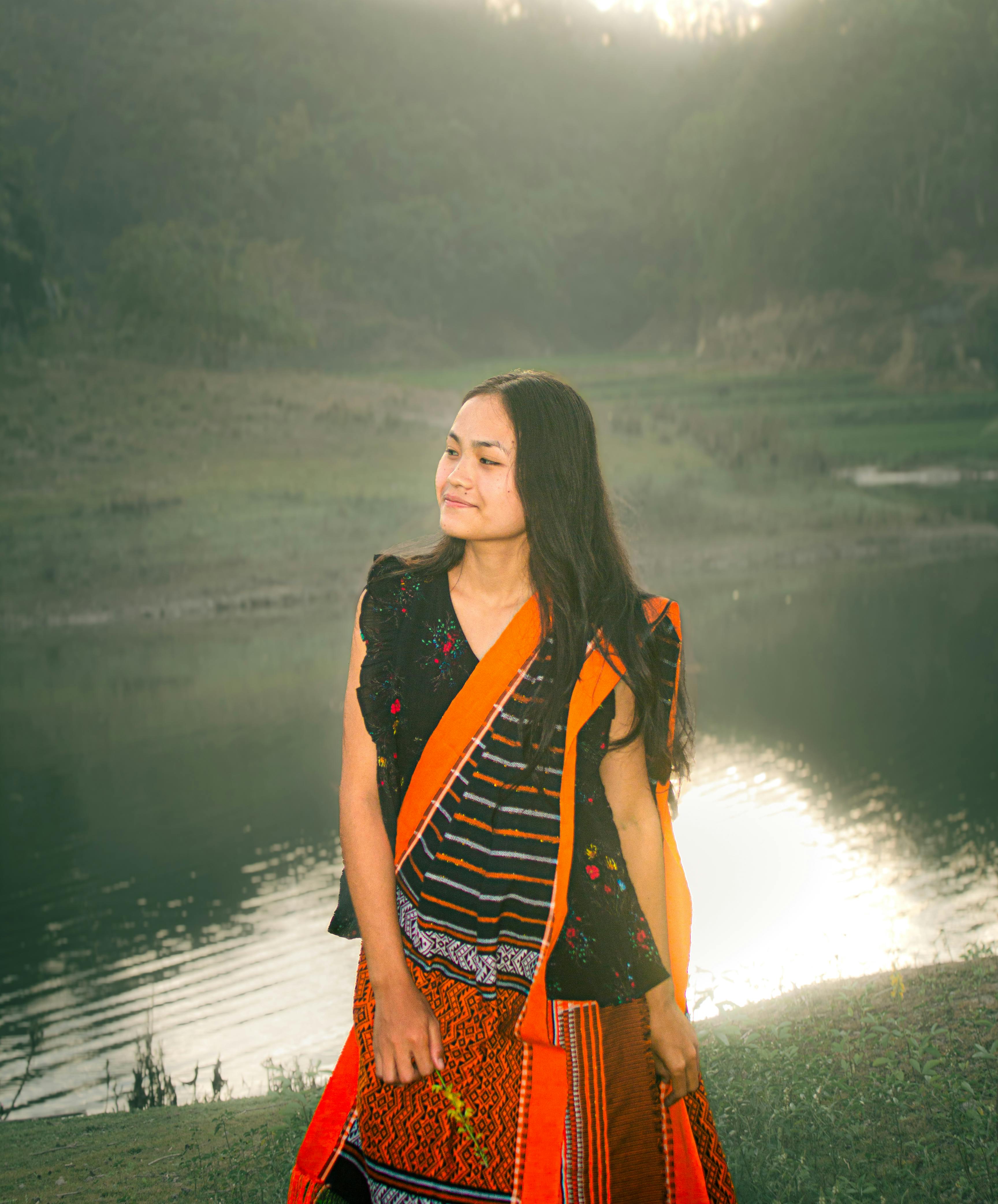 A serene woman in vibrant traditional clothing stands by a tranquil lake at sunset in Rangamati, Bangladesh.
