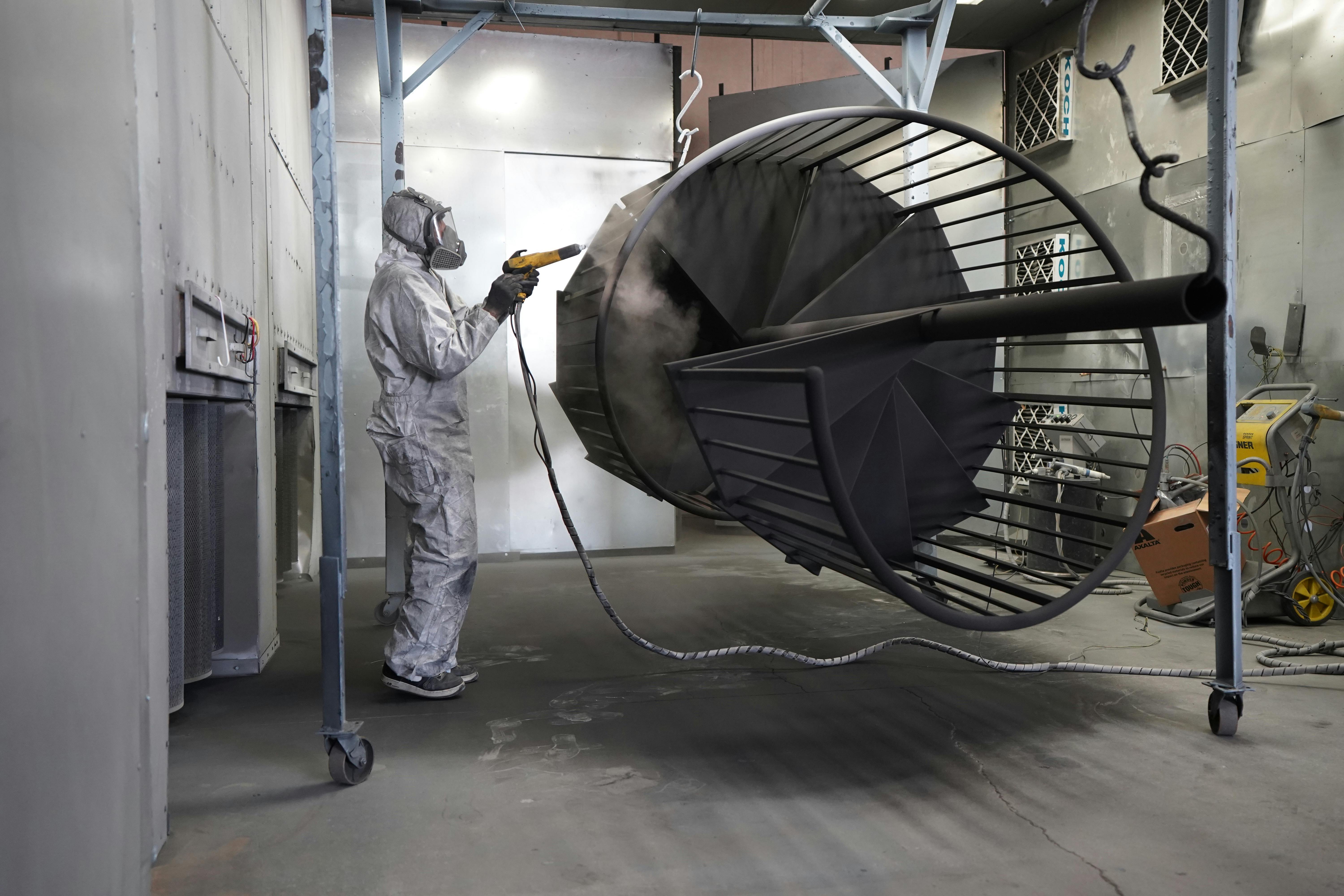 Worker in protective gear spray painting a large metal structure in an industrial facility.