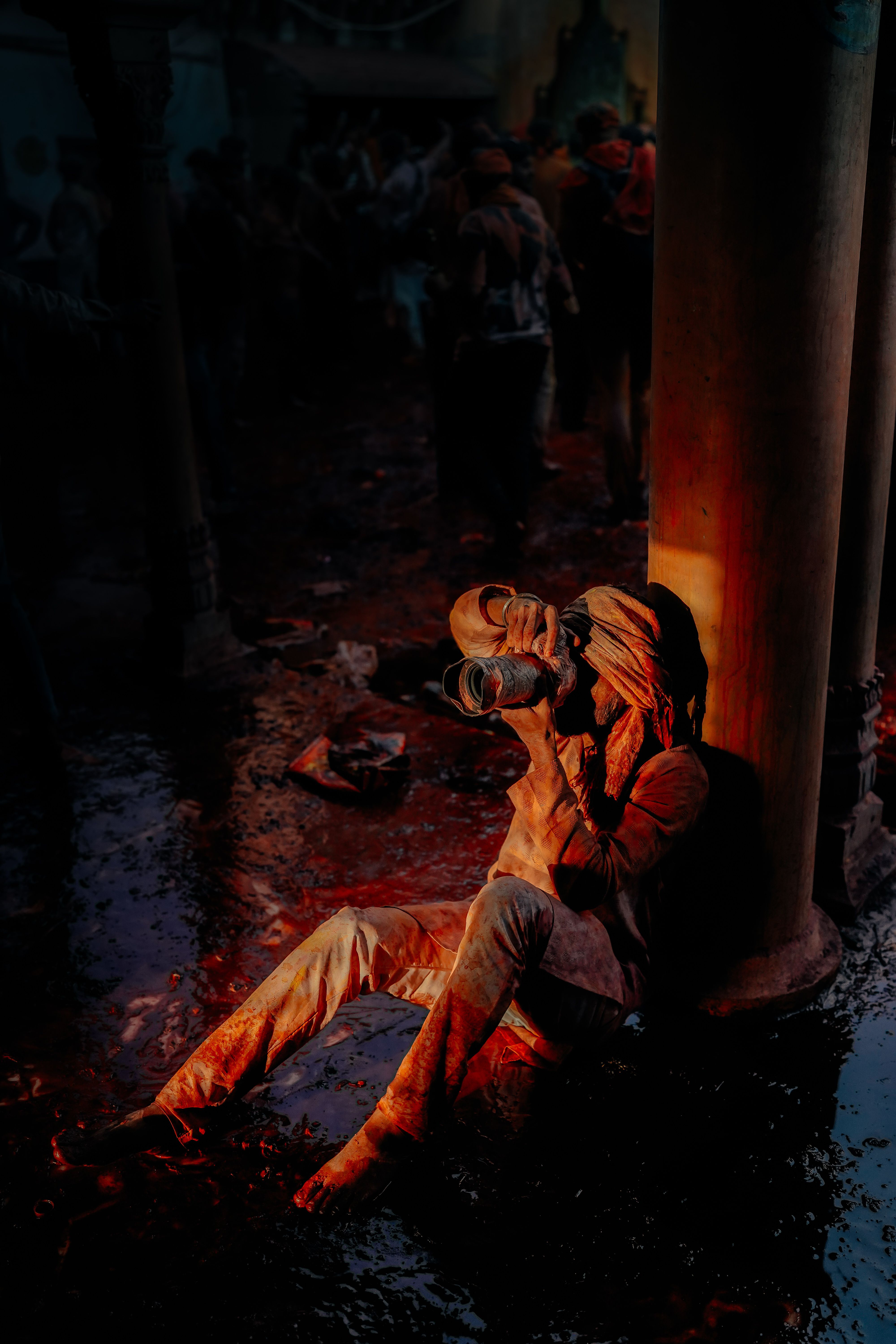 A person covered in colorful powder relaxes against a pillar during Holi.
