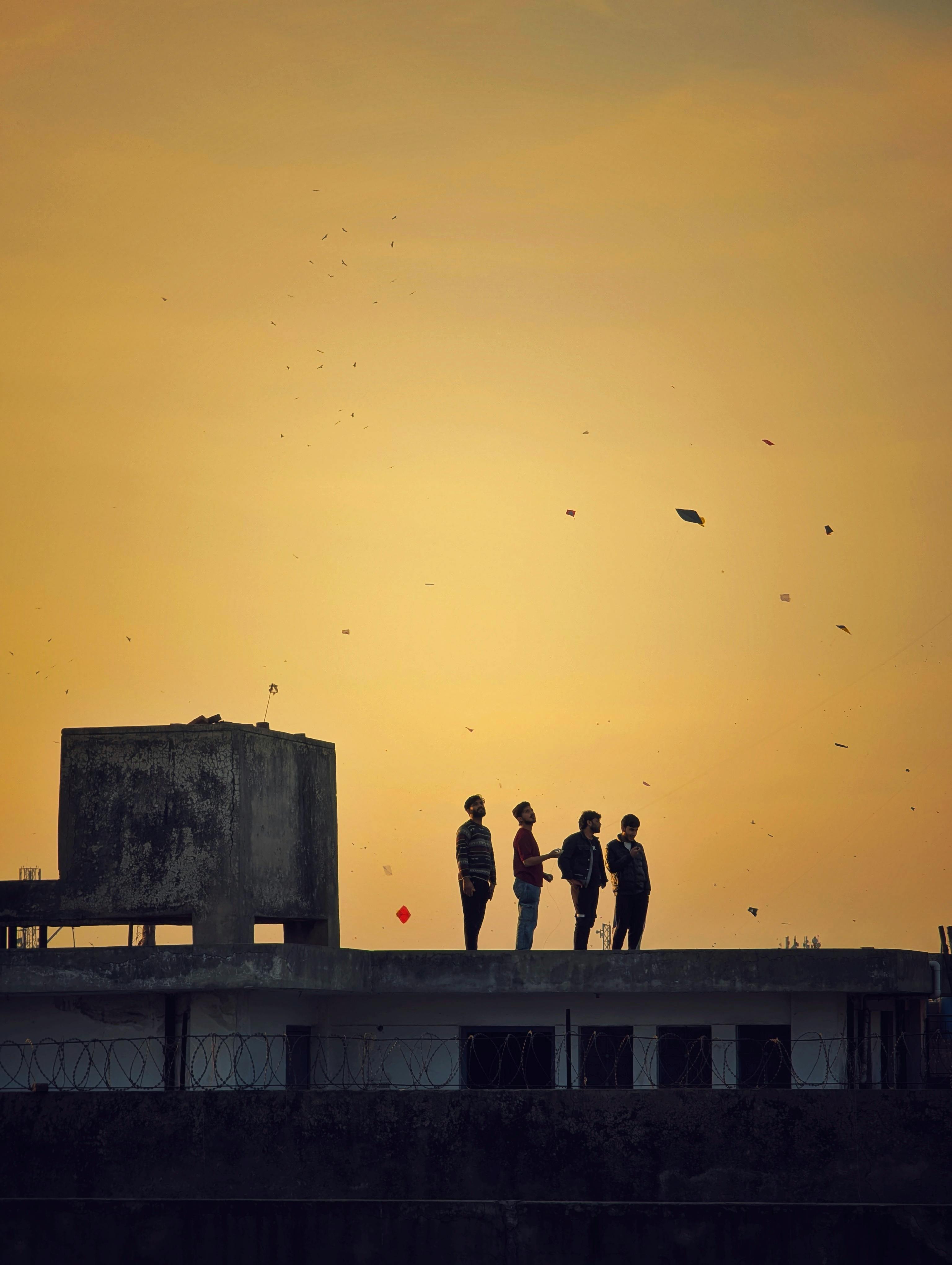 ฟรี ภาพเงาของคนสี่คนตัดกับท้องฟ้าในยามพระอาทิตย์ตกดิน กำลังเล่นว่าวอยู่บนดาดฟ้า สื่อถึงการผจญภัยของวัยรุ่น คลังภาพถ่าย