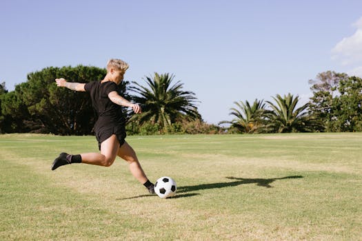A woman soccer player kicking a ball on a grassy field during a sunny day outdoors.