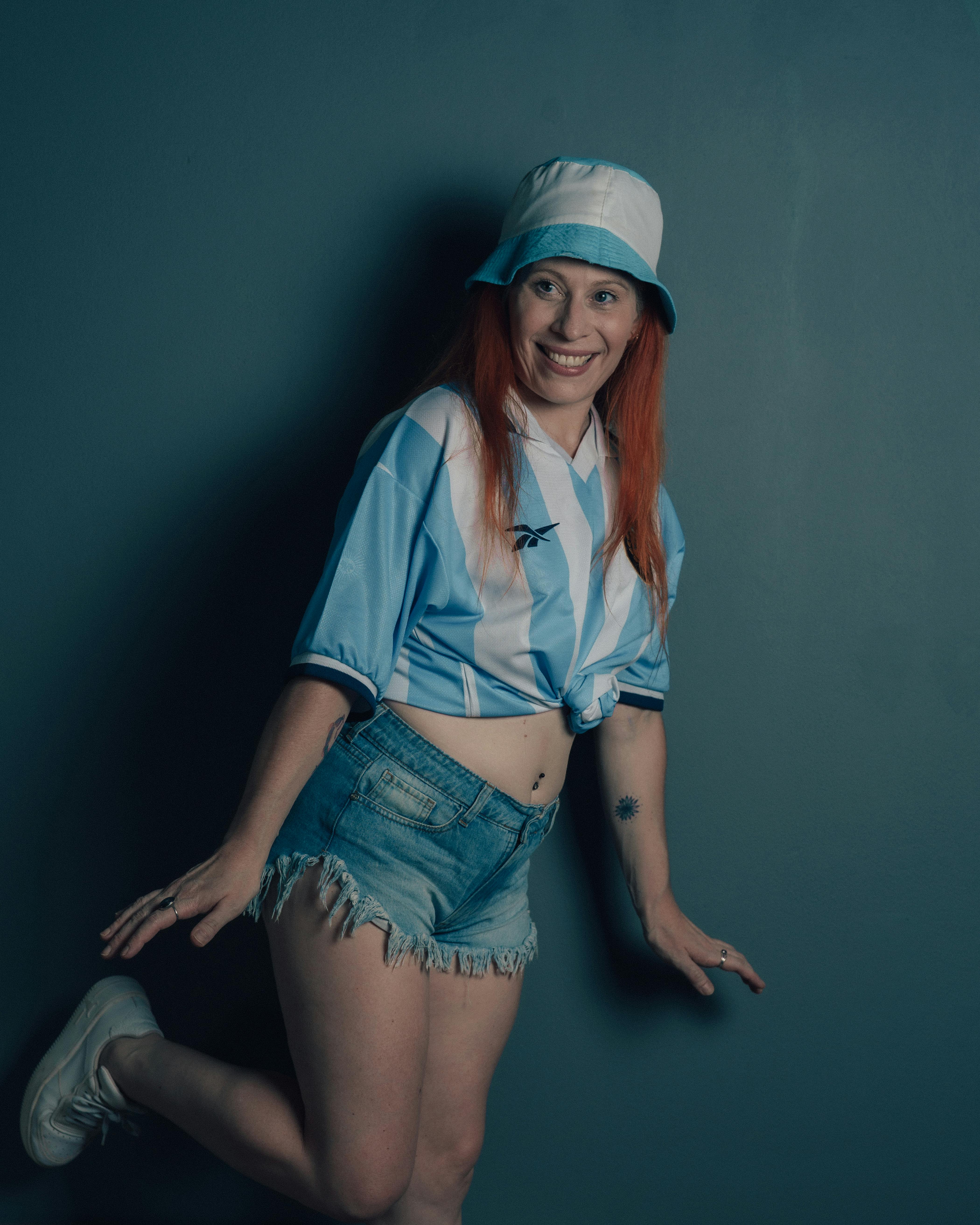 Gratis Donna felice in posa con maglia da calcio, pantaloncini di jeans e cappello da pescatore. Foto a disposizione