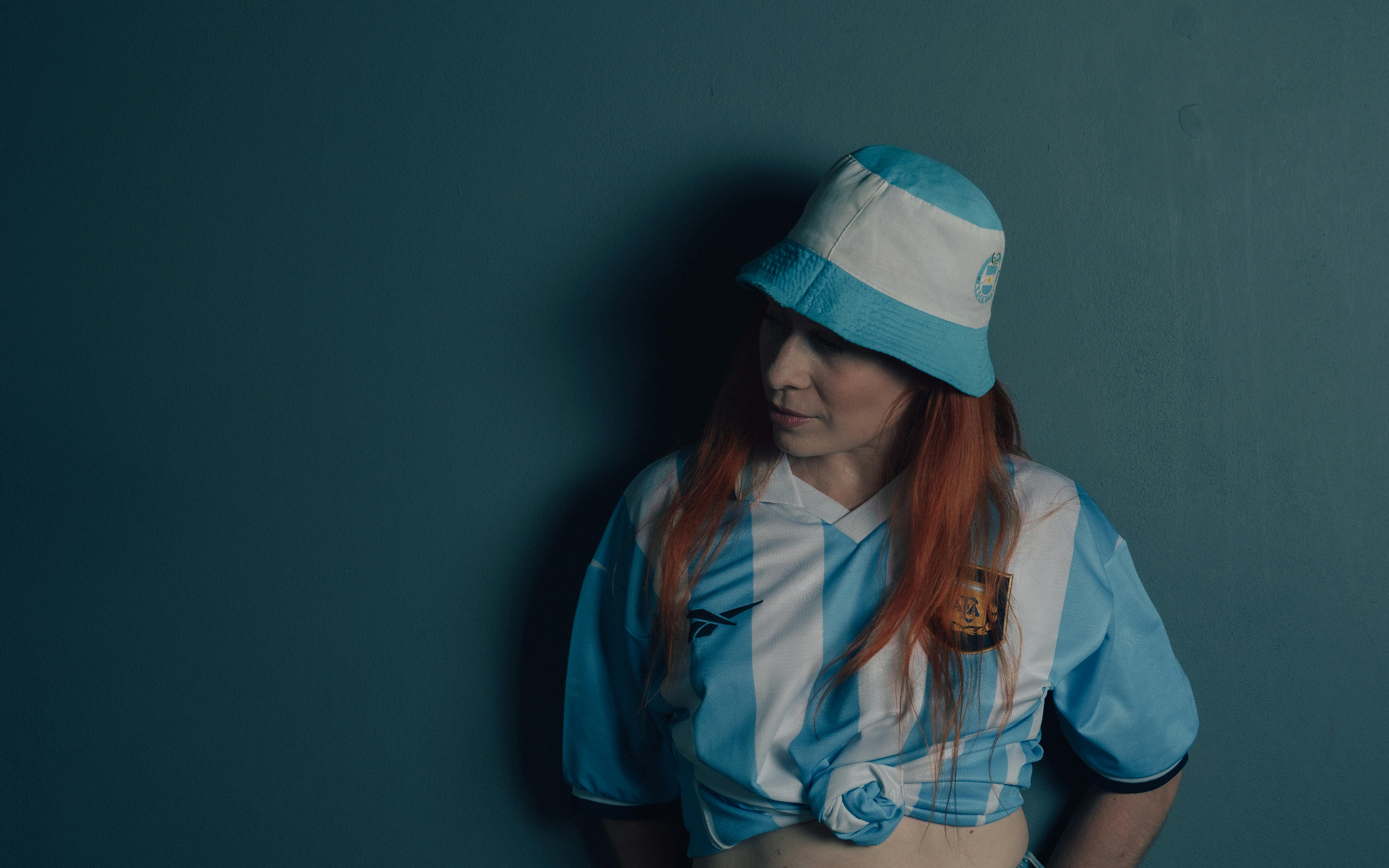 bezplatná Dospělá žena v dresu a čepici argentinského fotbalového týmu, stojící uvnitř s náladovým pozadím. Základová fotografie