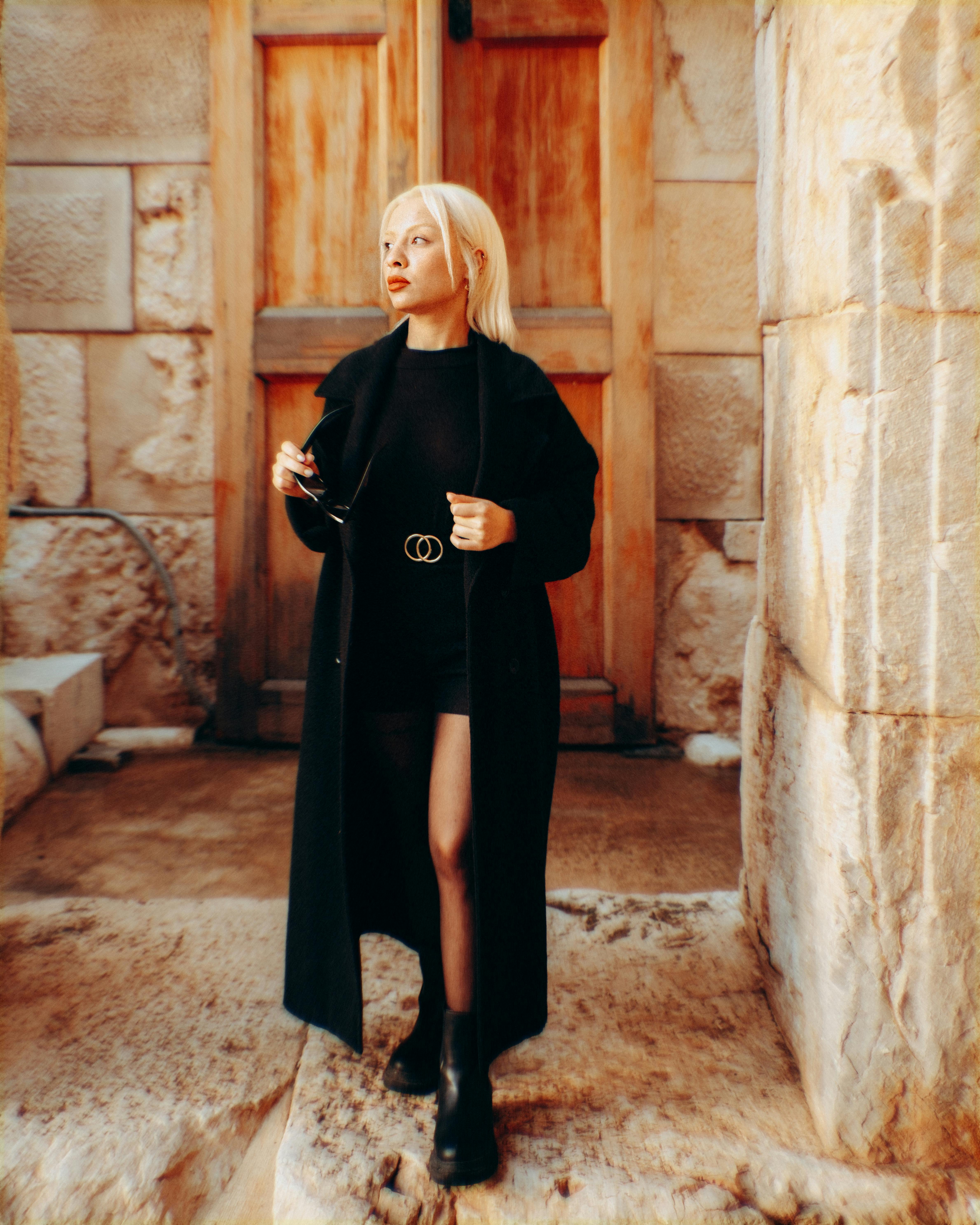 ฟรี หญิงสาวทันสมัยในชุดสีดำยืนอย่างมั่นใจอยู่หน้าประตูที่มีพื้นผิวขรุขระแบบชนบท คลังภาพถ่าย