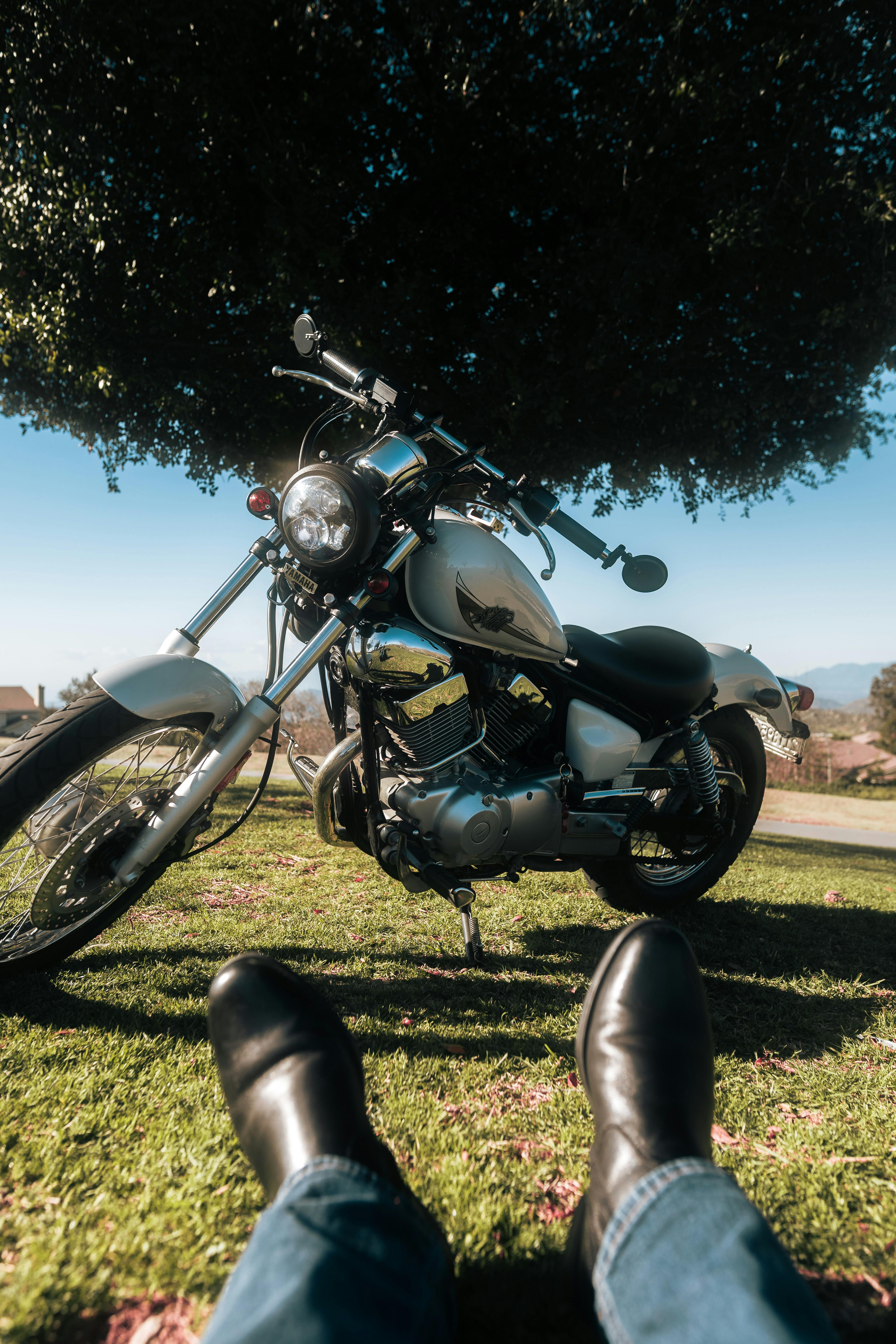 ฟรี รถจักรยานยนต์จอดอยู่ใต้ต้นไม้ โดยมีรองเท้าบูทอยู่ด้านหน้า ในวันที่แดดจ้า คลังภาพถ่าย