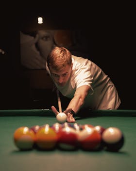 A young man intensely focuses on making a billiard shot in a dimly lit room.