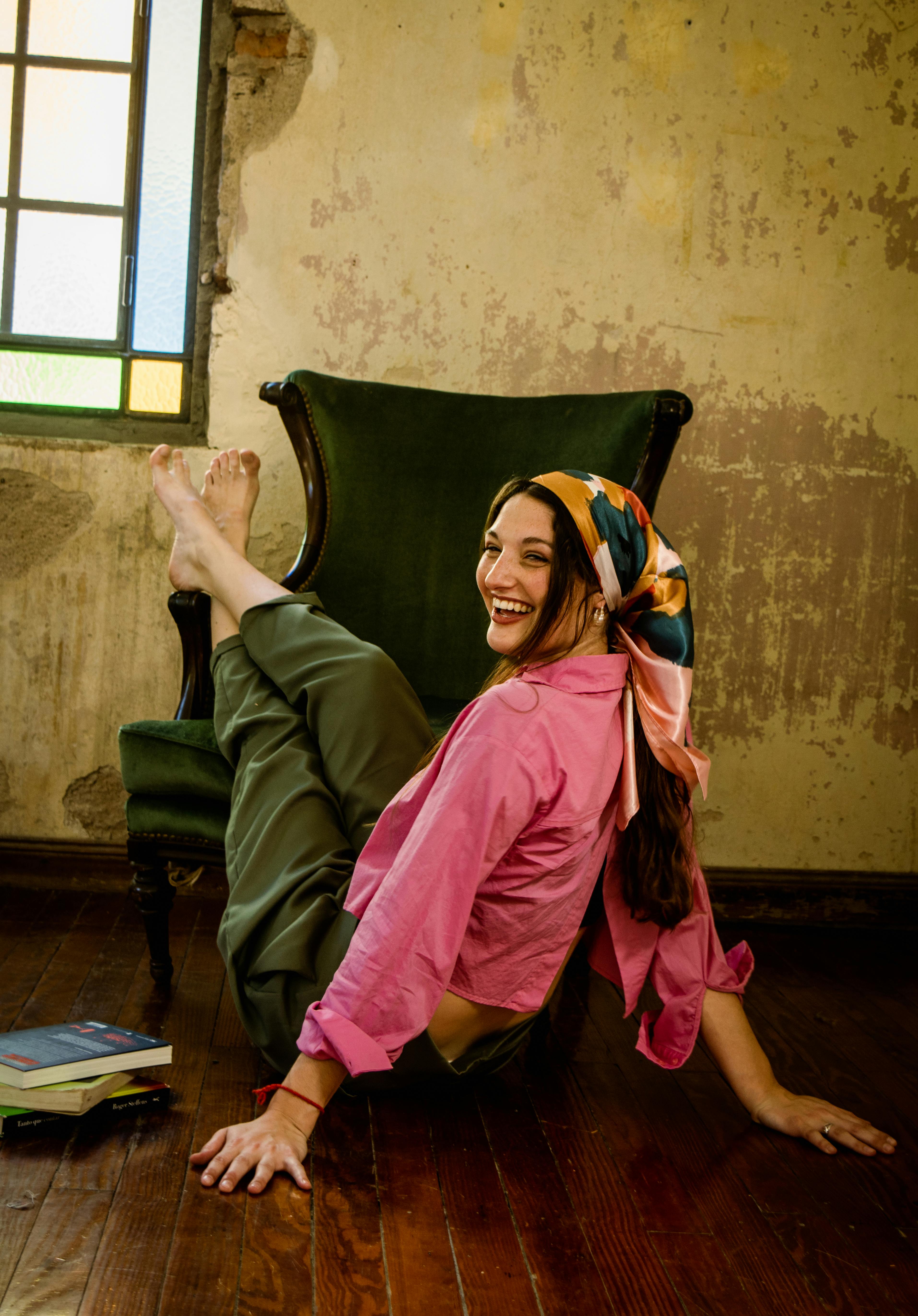 A cheerful woman lounges barefoot with a colorful headscarf and pink shirt indoors.
