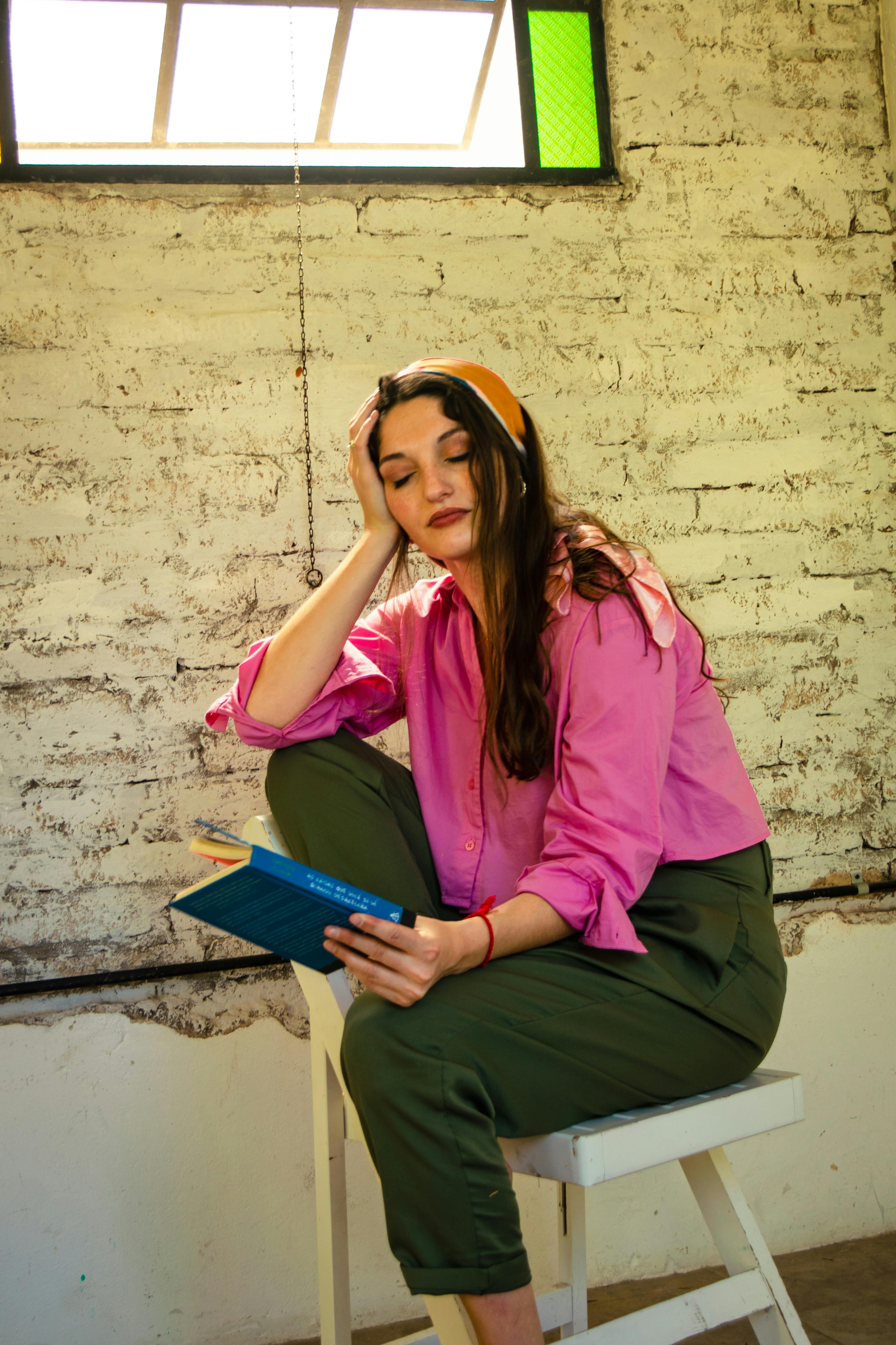 bezplatná Mladá žena v barevném oblečení čte knihu uvnitř u rustikální cihlové zdi. Základová fotografie