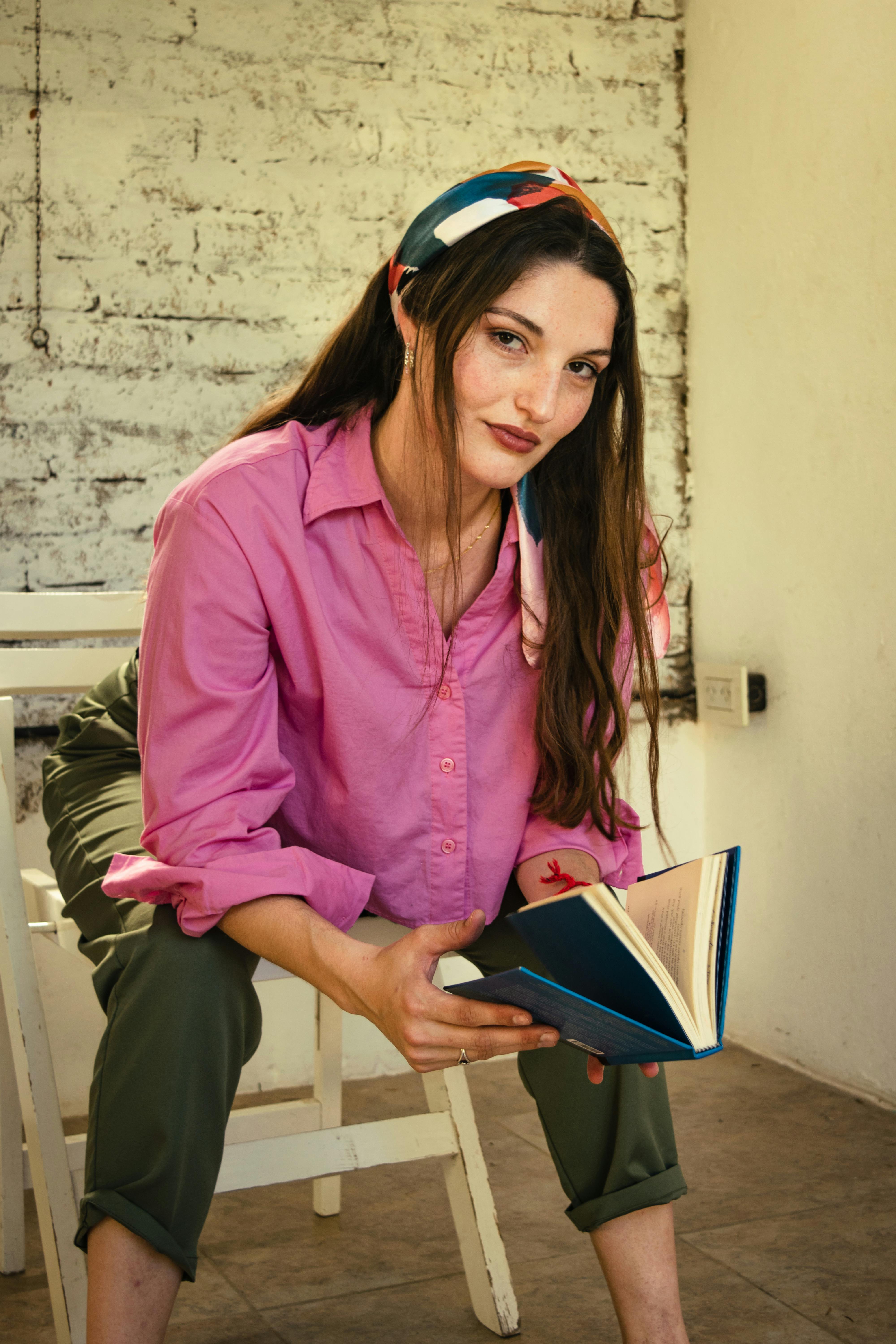 Gratis Una donna con una camicia rosa legge un libro in casa, con un'eleganza disinvolta. Foto a disposizione