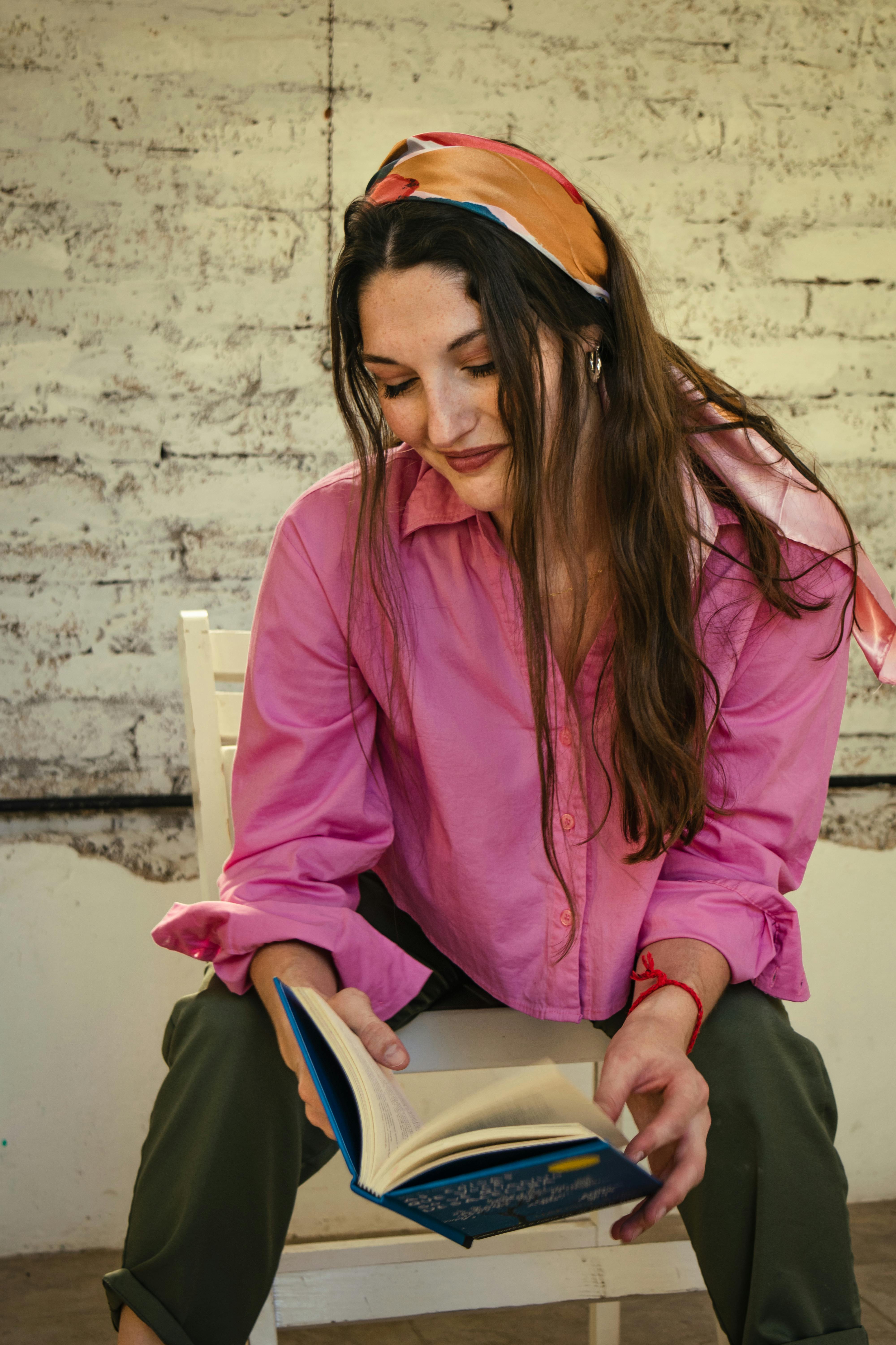 Free A young woman in a pink blouse sits on a chair reading a book against a rustic wall. Stock Photo