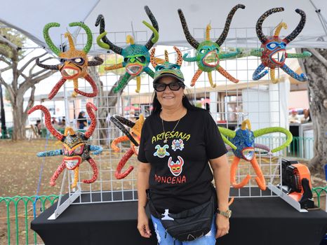 A woman showcases vibrant, colorful artisan masks at an outdoor market booth.