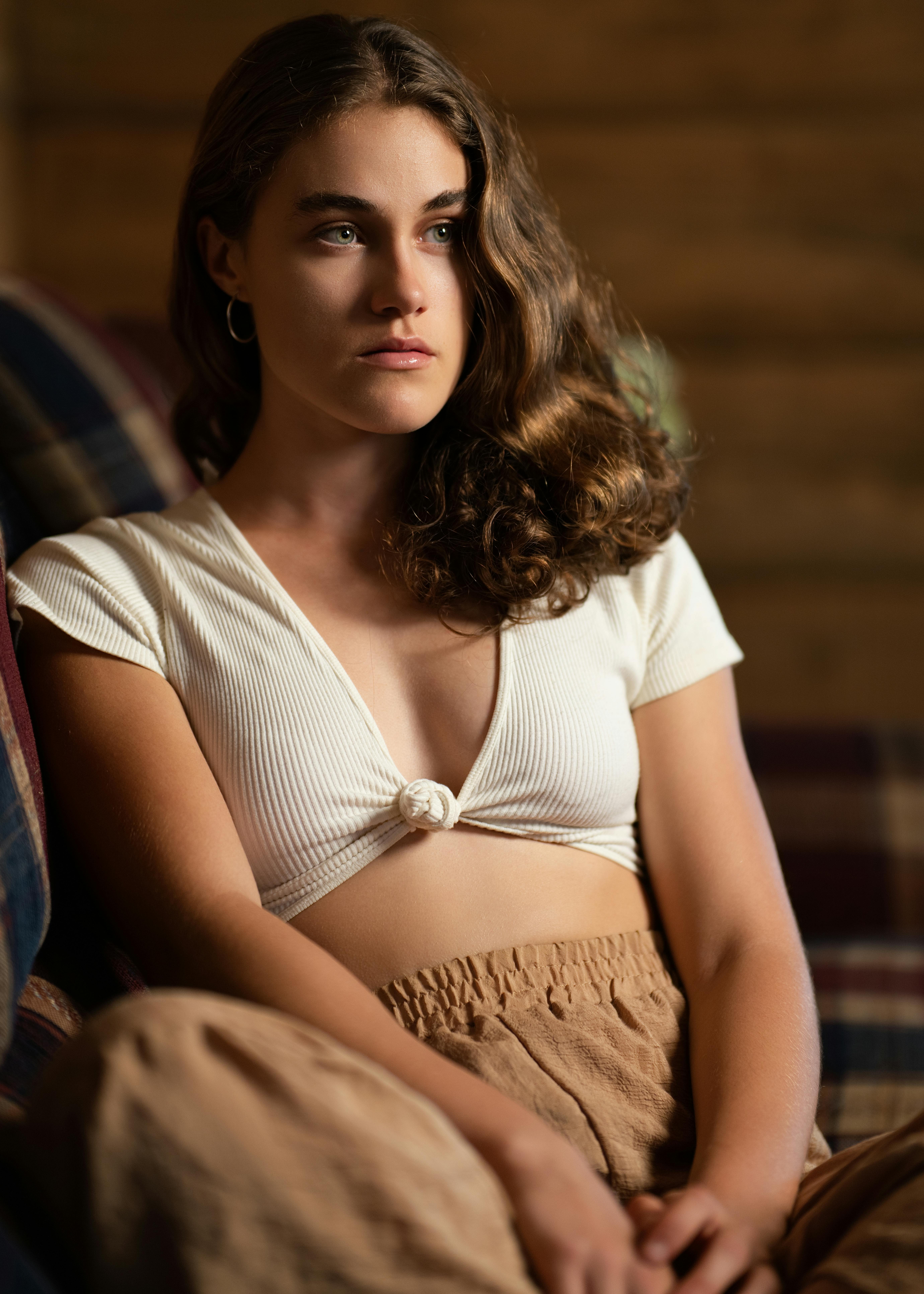 Gratis Una giovane donna con i capelli ricci seduta in un interno, immortalata in una luce calda e soffusa, che crea un'atmosfera riflessiva. Foto a disposizione