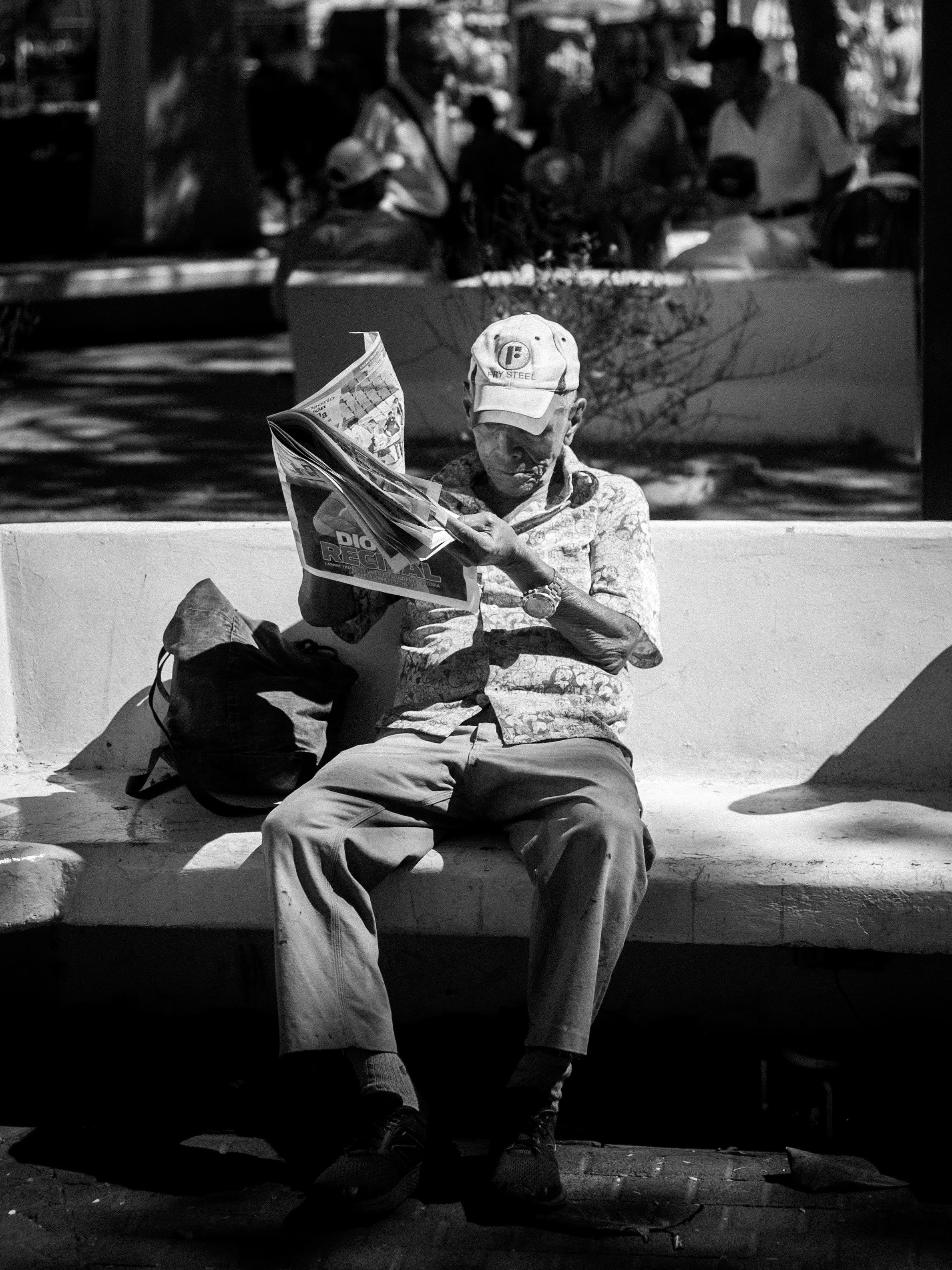 Ücretsiz Park bankında oturmuş, gazete okumaya dalmış yaşlı bir adamın siyah beyaz fotoğrafı. Stok Fotoğraflar