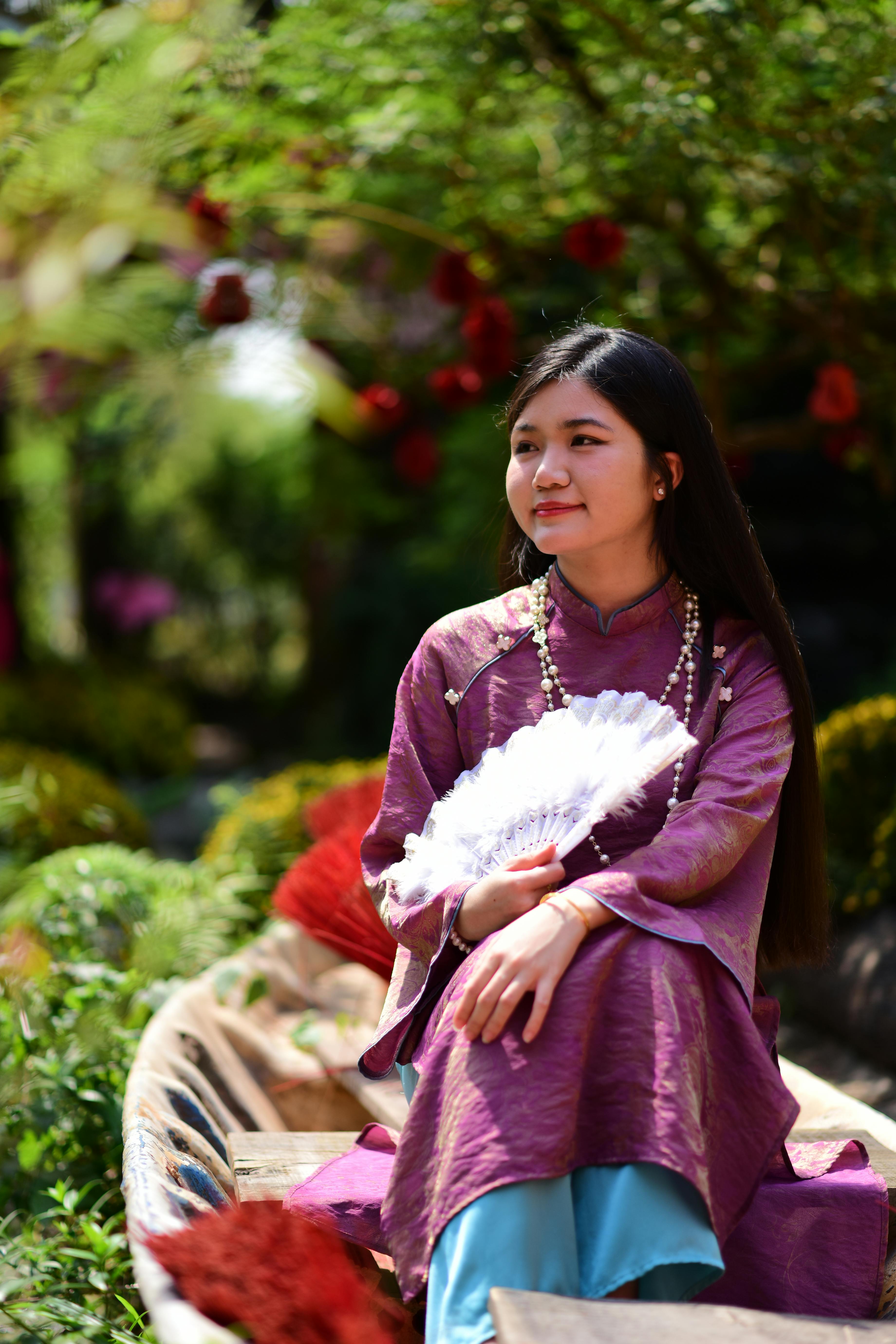 Gratis Una giovane donna in abiti tradizionali con un ventaglio in mano, seduta all'aperto in un vivace giardino. Foto a disposizione