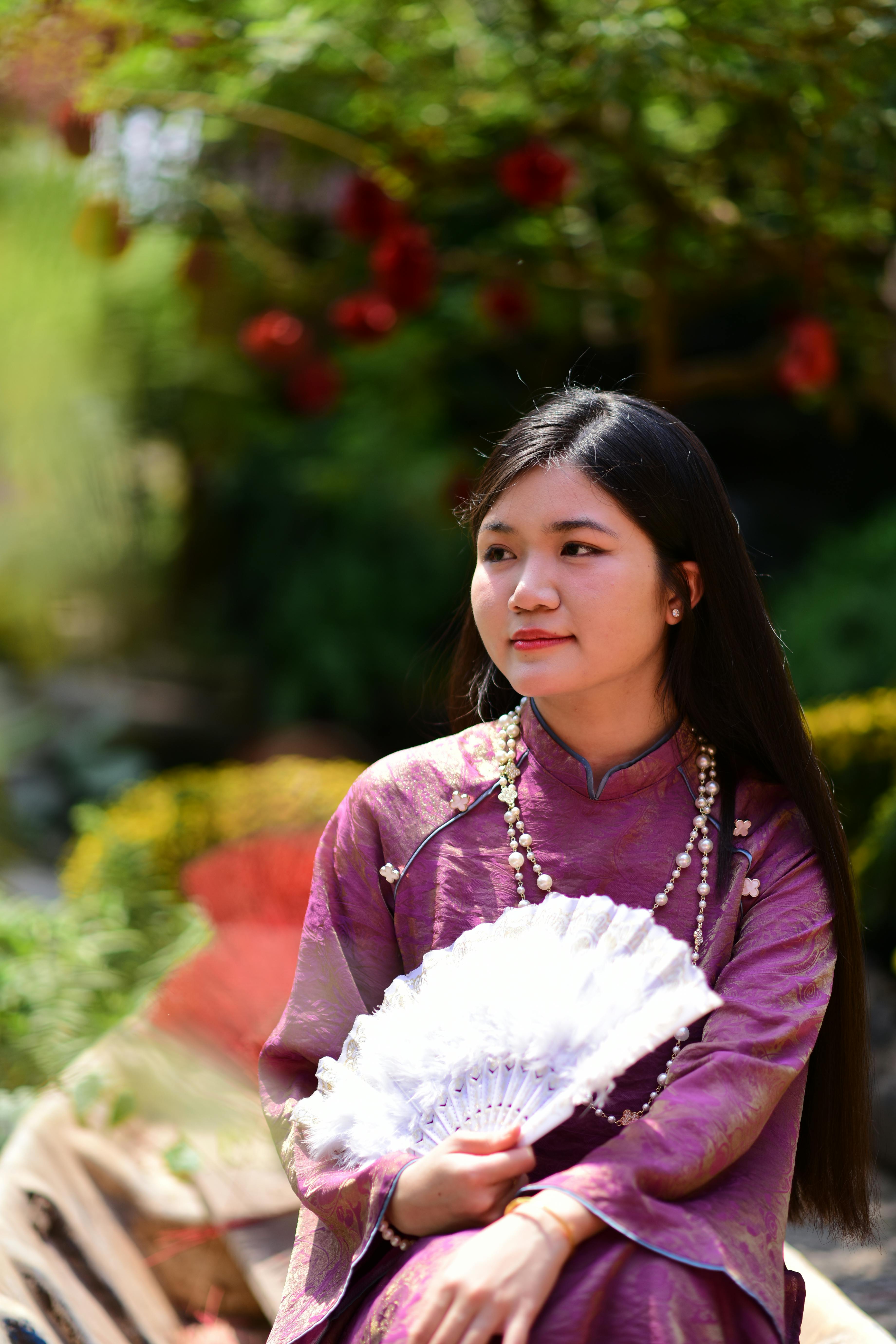 Ilmainen Rauhallinen nuori nainen perinteisessä silkkimekossa nauttii aurinkoisesta päivästä ulkona valkoisen viuhkan kanssa. Kuvapankkikuva