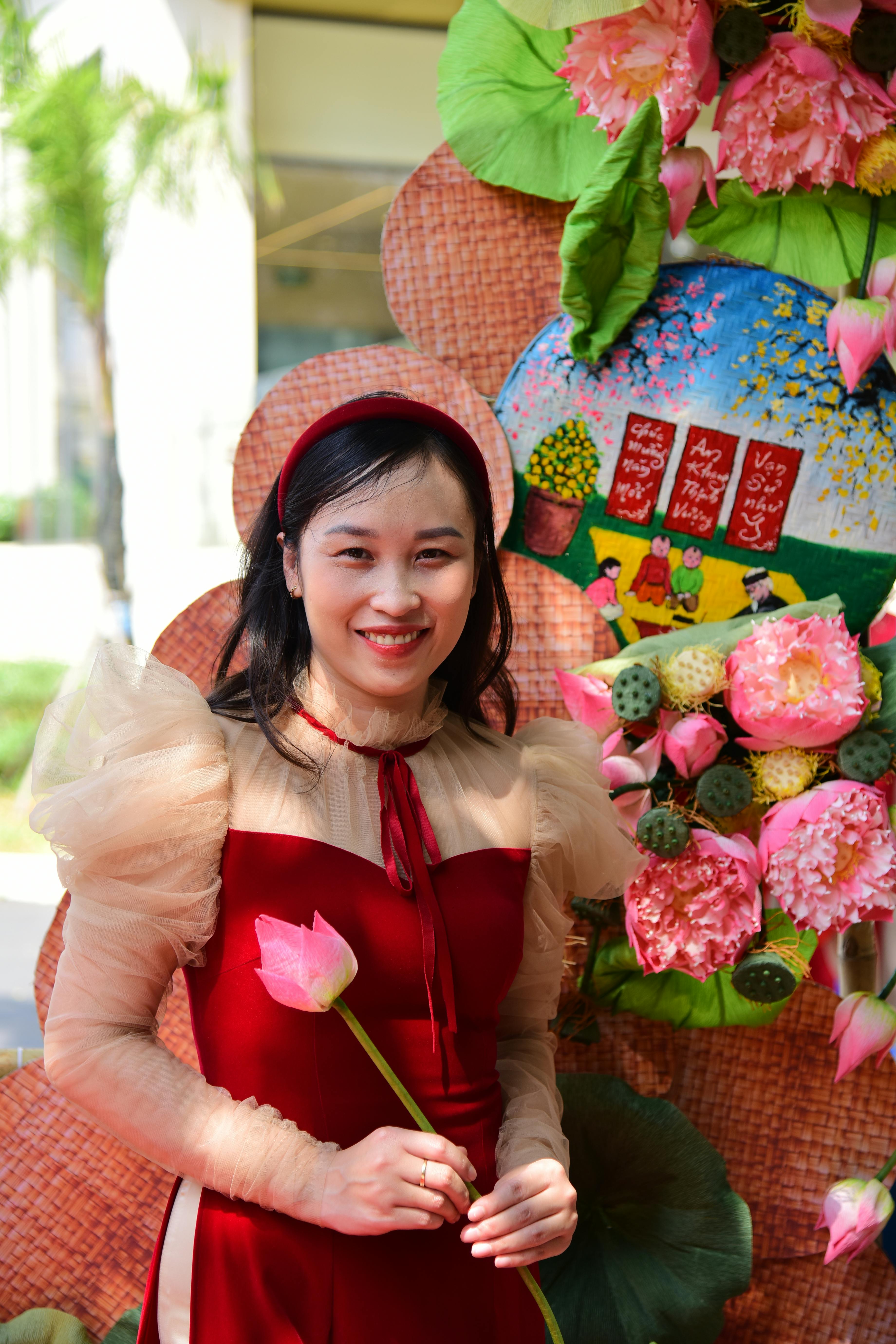 Ilmainen Hymyilevä nainen elegantissa punaisessa mekossa pitelee lootuskukka juhlavan kukkaisen taustan edessä. Kuvapankkikuva