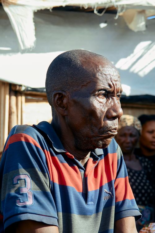 Free A contemplative elderly man in a striped shirt stands outdoors with people in the background. Stock Photo