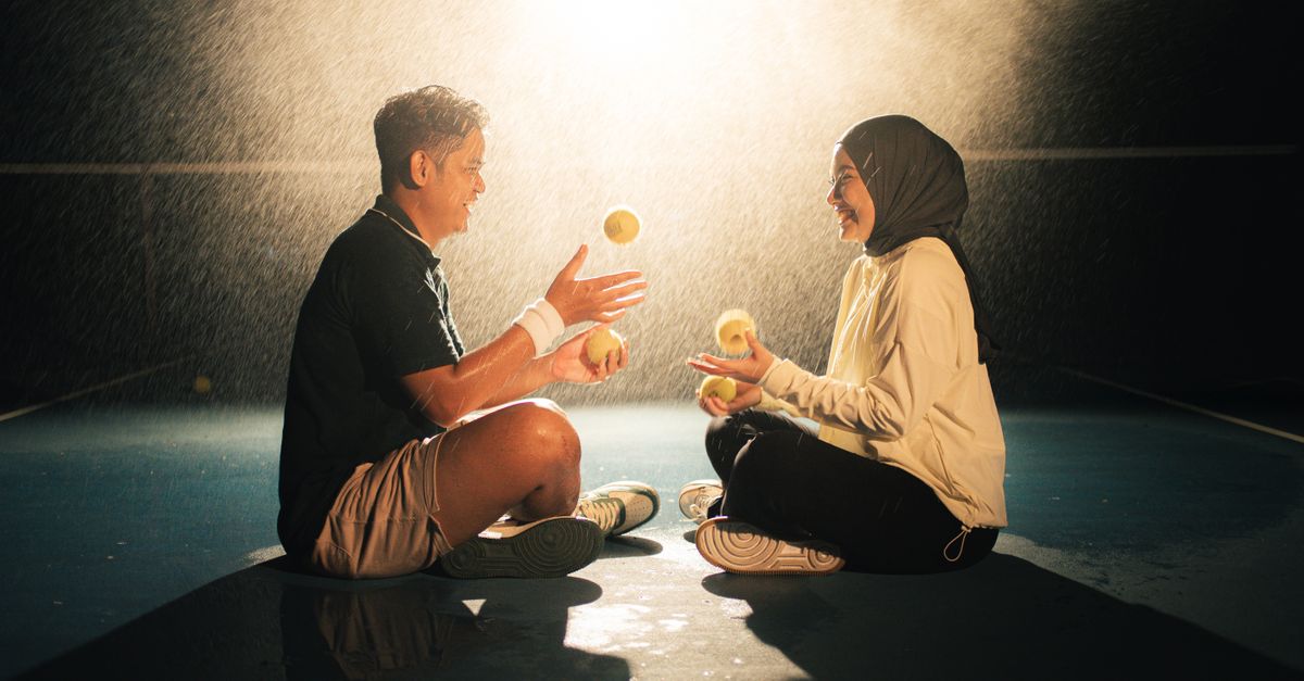 A joyful couple juggling tennis balls on a court at night under dramatic lighting.