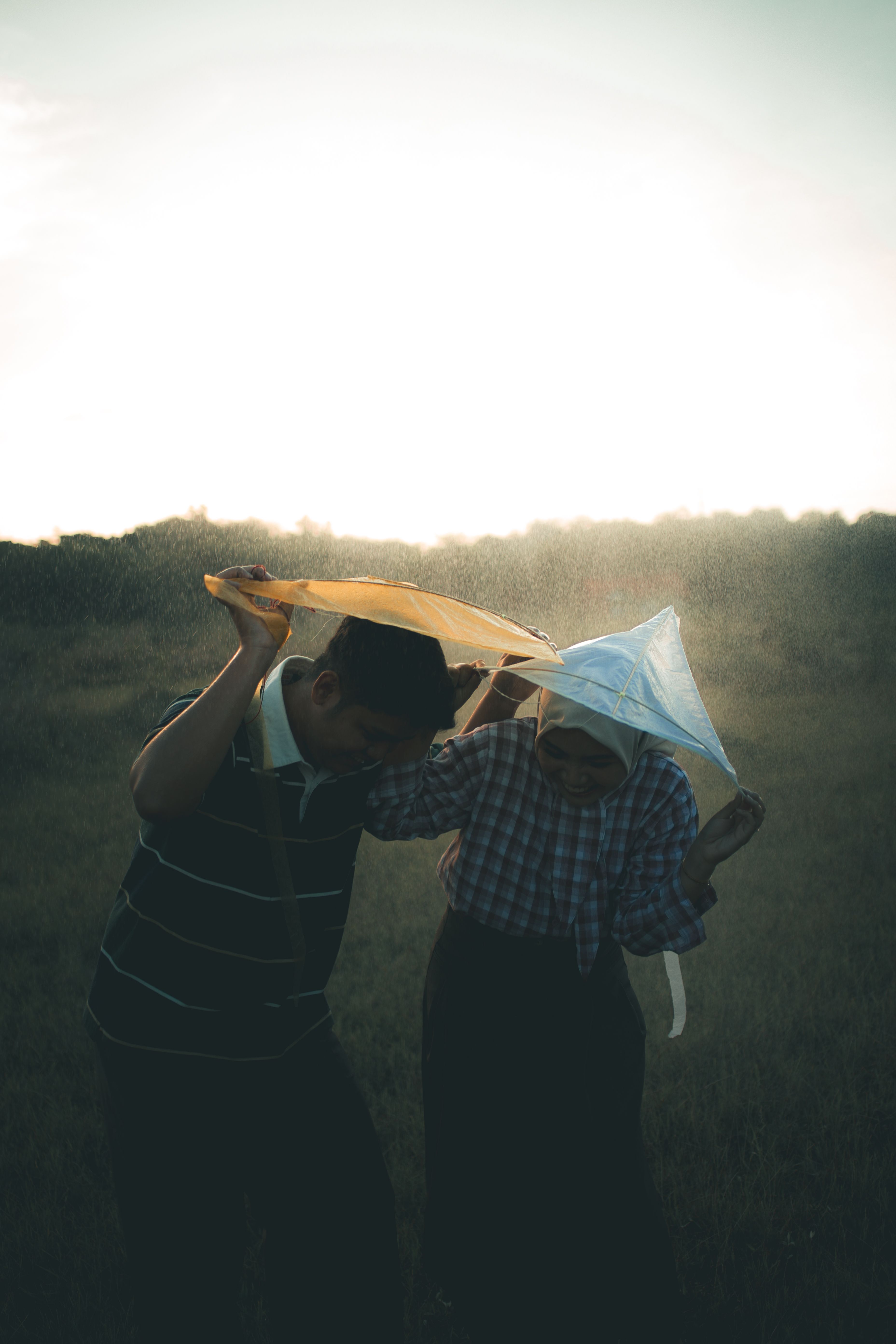 An engaged couple in Indonesia enjoys a playful moment under umbrellas in a lush countryside setting.