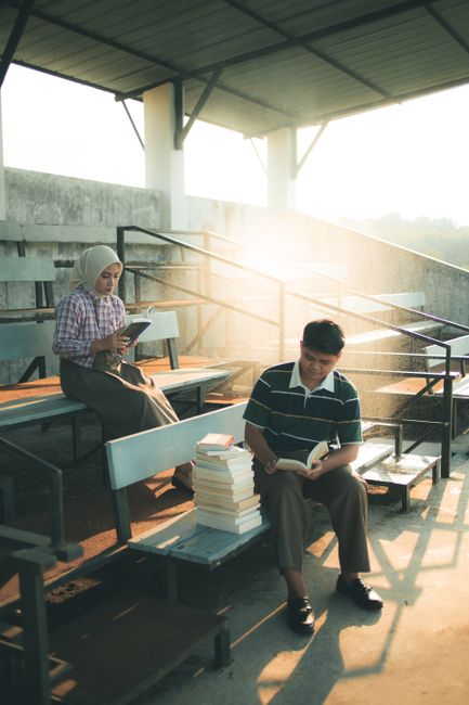 Two students study on sunlit bleachers in Indonesia, surrounded by books.