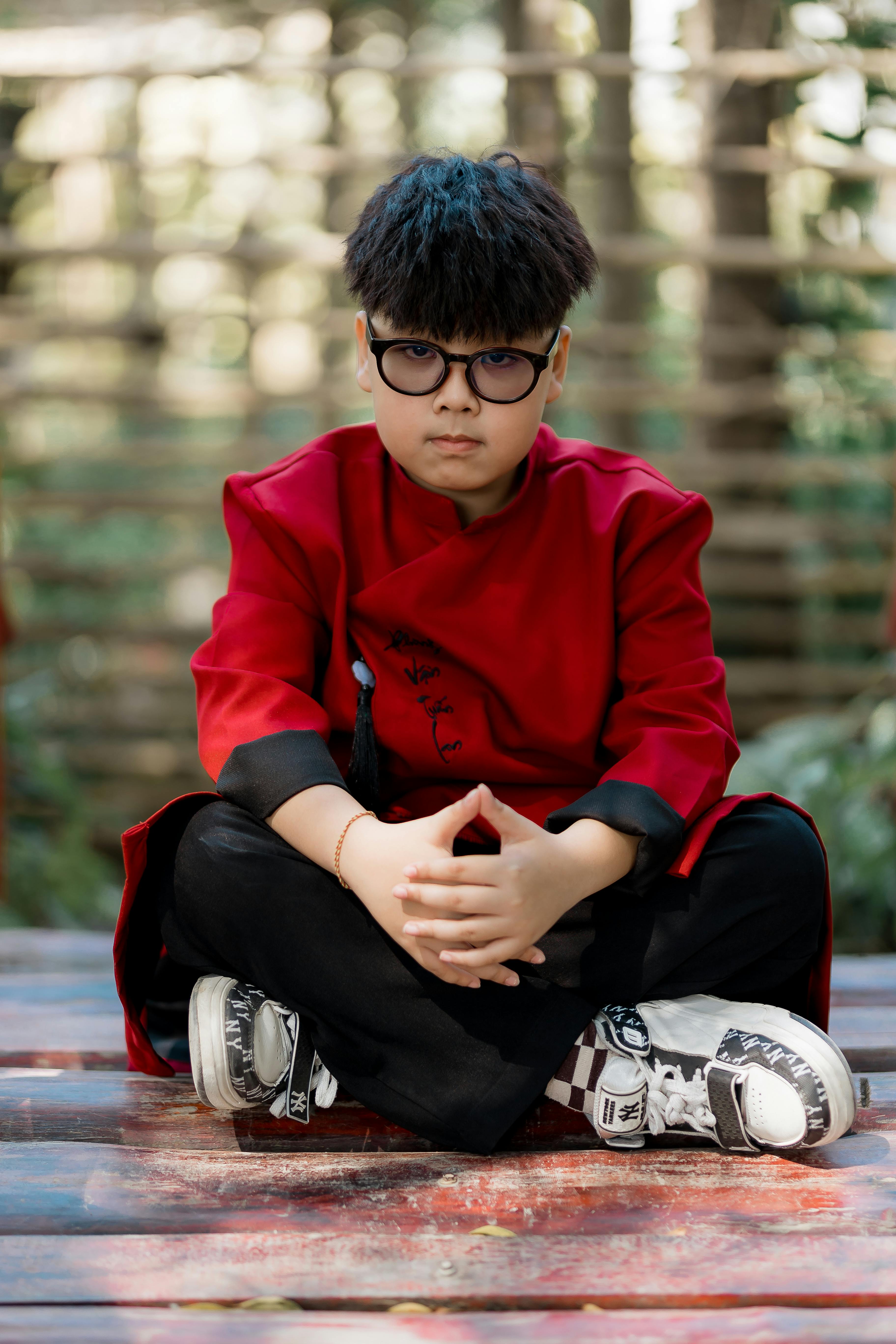 ingyenes Fiatal fiú ül törökülésben a szabadban, piros kabátban és szemüvegben. Stockfotó