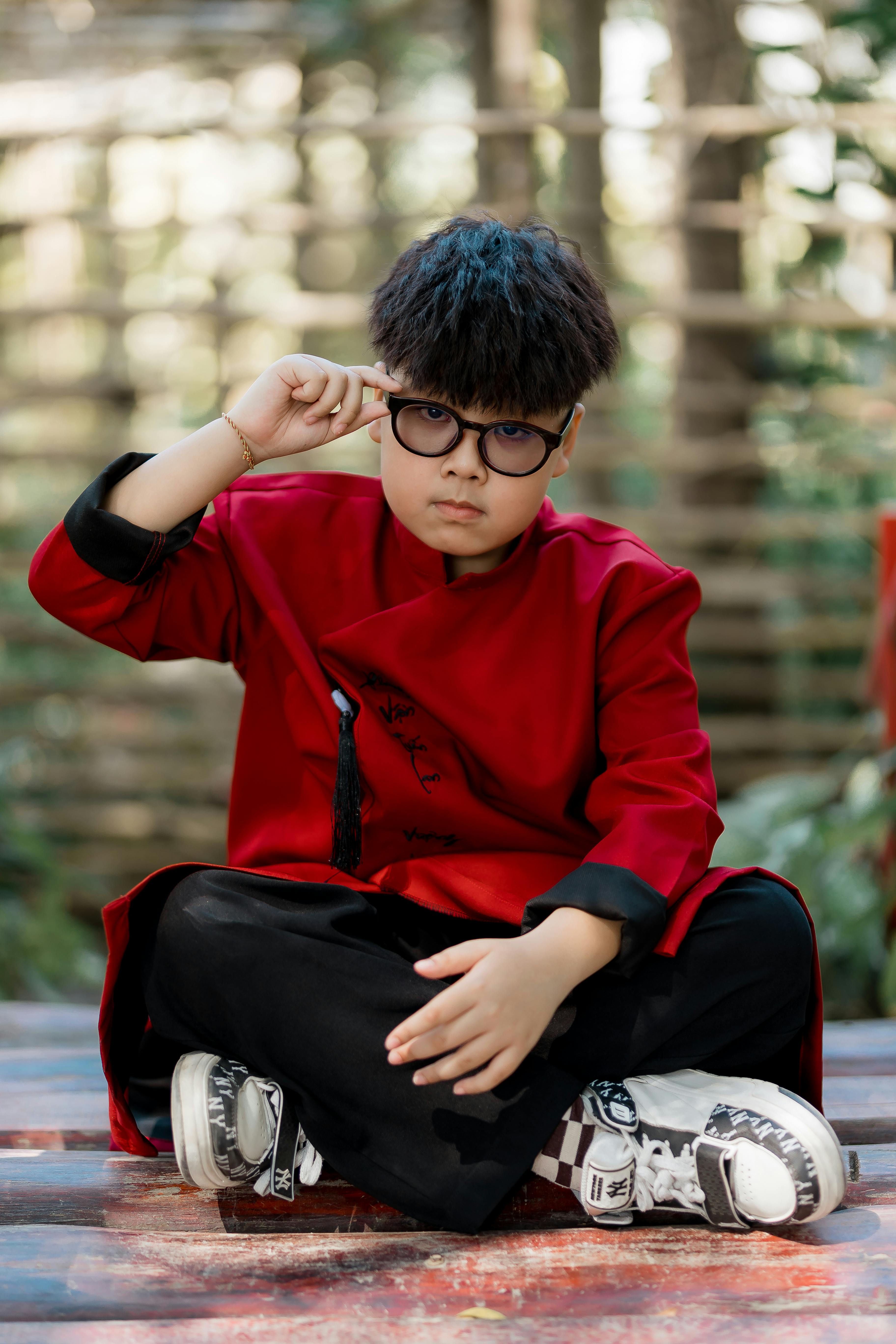 Bezpłatne Chłopiec siedzi na zewnątrz w tradycyjnym, czerwonym stroju, w okularach i swobodnie pozuje. Zdjęcie z galerii