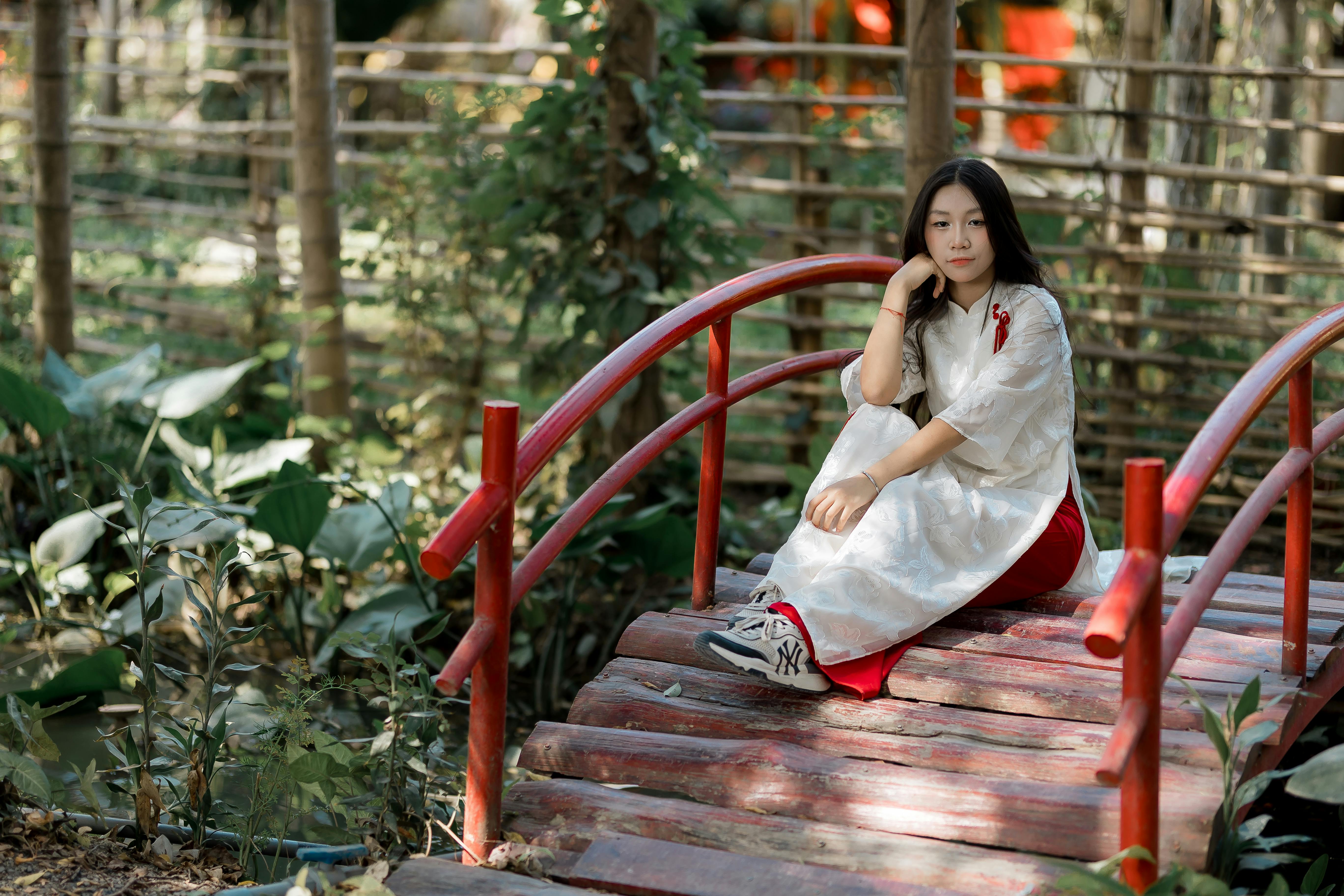 Free Young woman in traditional attire sitting on a red wooden bridge in a lush garden. Stock Photo