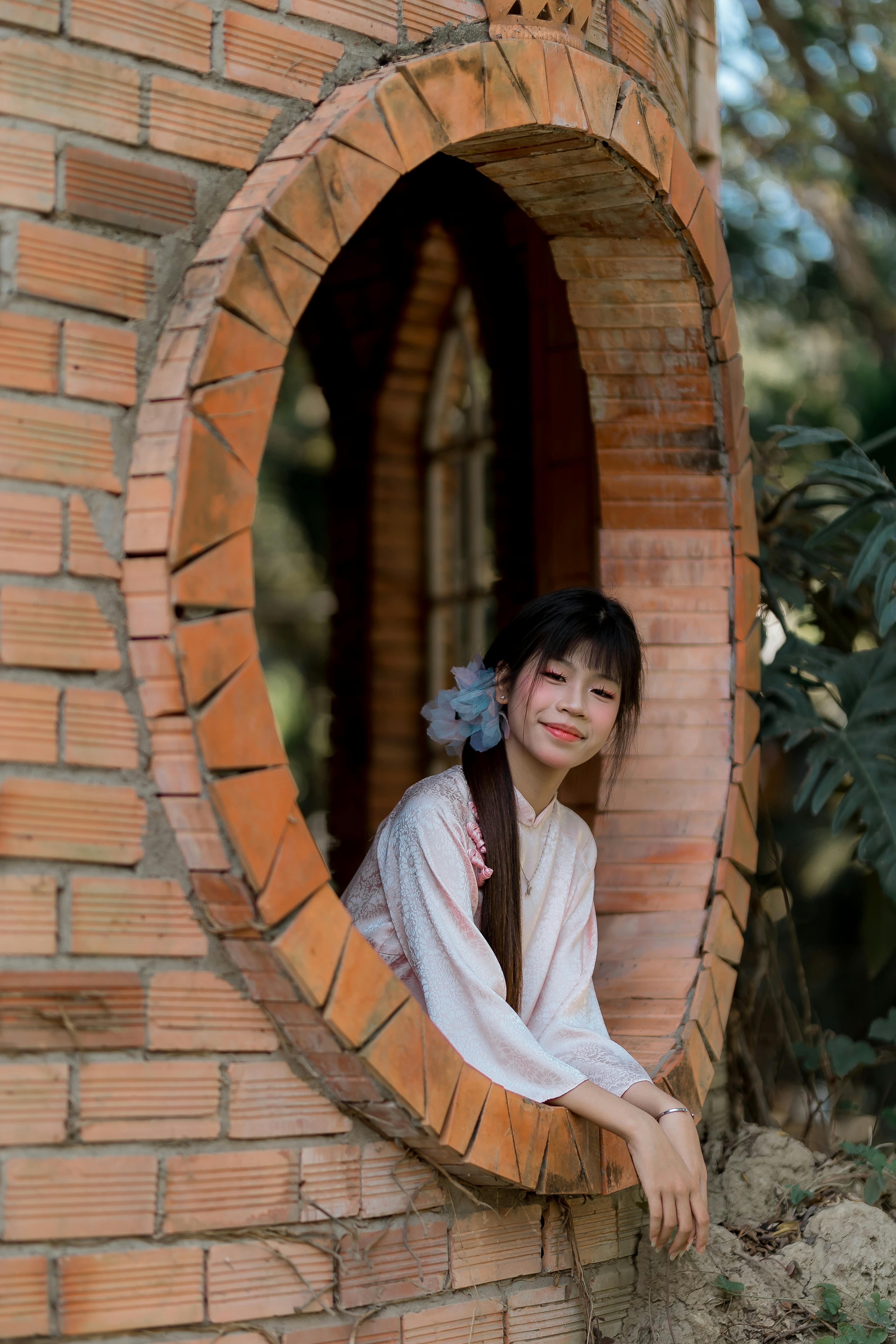 Ilmainen Perinteisiin vaatteisiin pukeutunut nuori nainen hymyilee pyöreästä tiili-ikkunasta herättäen lämpimiä, luonnollisia tunnelmia. Kuvapankkikuva