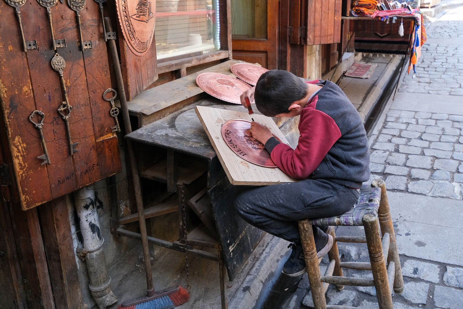 A young boy skillfully engraving a metal plate in a traditional street setting, showcasing artisanal craftsmanship.