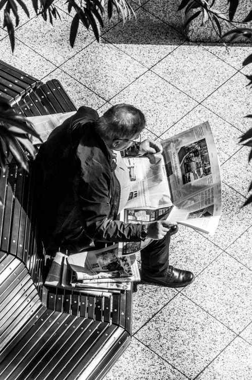 Free Aerial view of a man sitting on a bench reading a newspaper in a black and white urban setting. Stock Photo