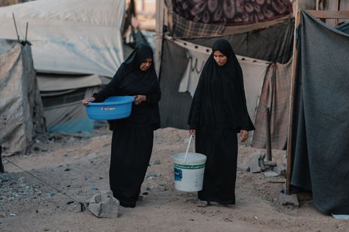 Free Two women in black hijabs carry water in a Gaza refugee camp, symbolizing resilience. Stock Photo