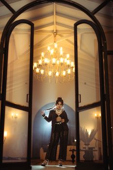 Stylish woman poses with chandelier backdrop inside elegant hall.