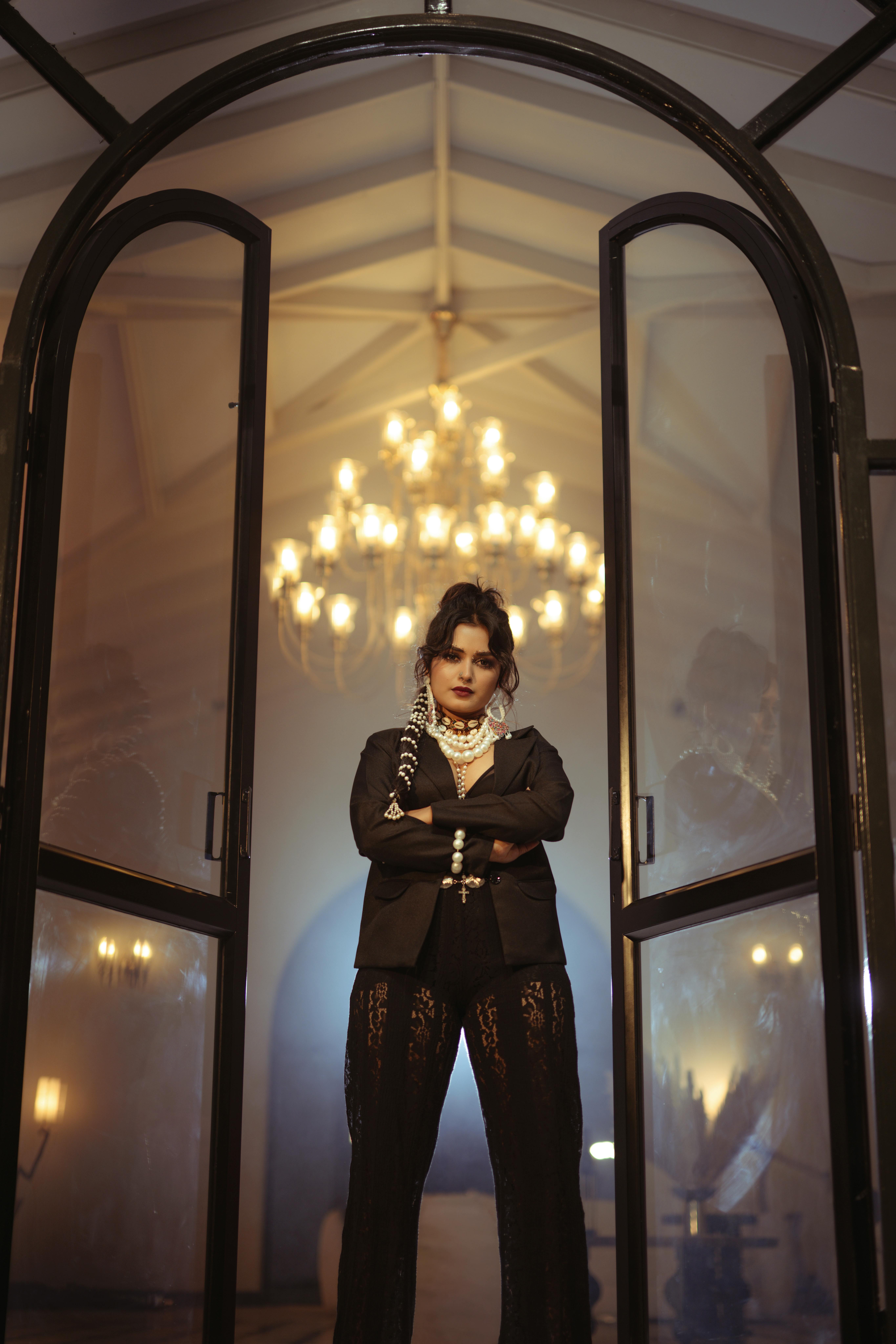 Ilmainen Hienostunut nainen poseeraa sisätiloissa kattokruunujen kanssa virallisessa ympäristössä, eleganssia huokuen. Kuvapankkikuva