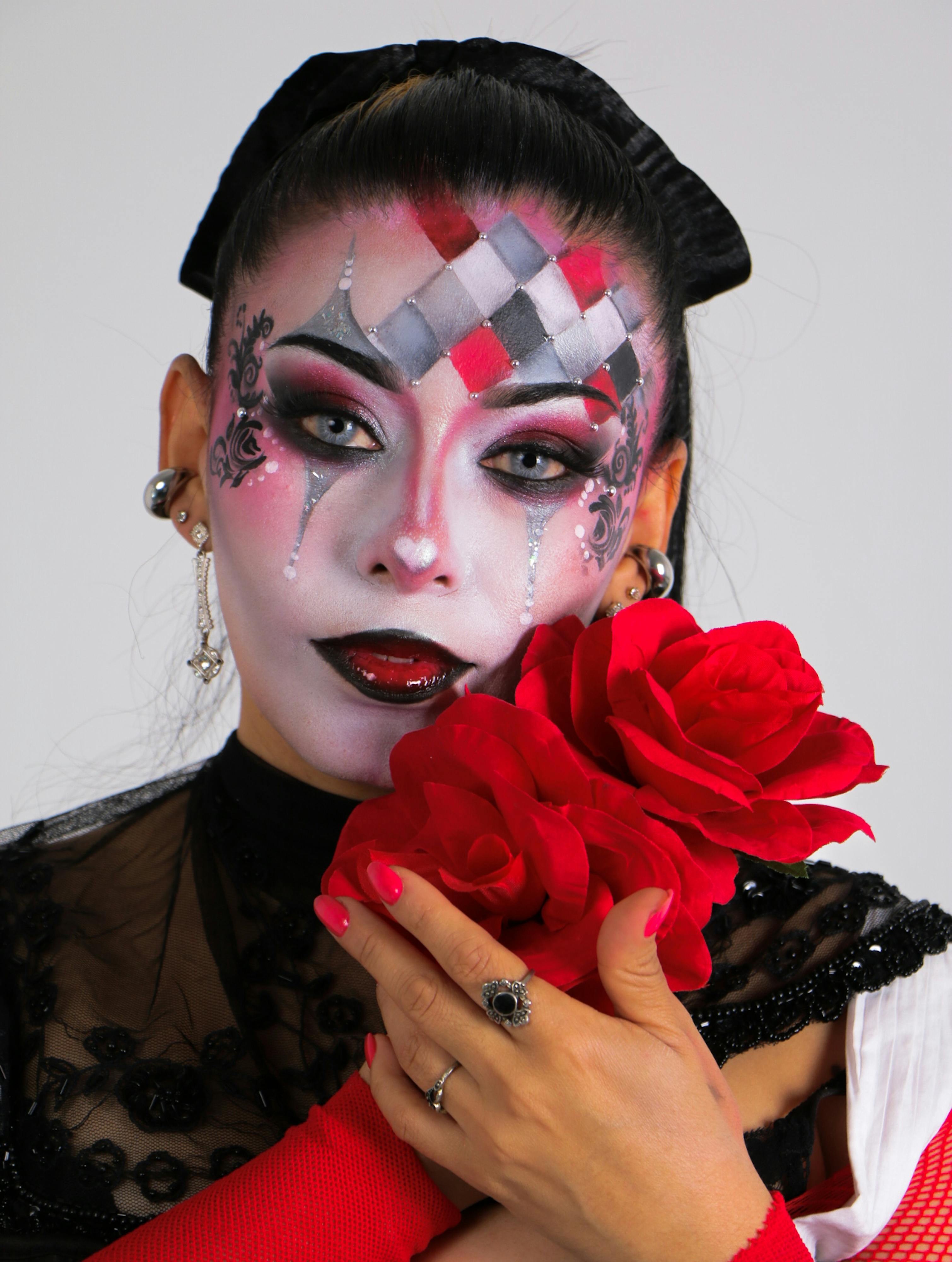 Ücretsiz Yüzüne soytarı makyajı yapılmış ve elinde kırmızı güller tutan, tiyatral bir zarafet yansıtan bir kadının canlı görüntüsü. Stok Fotoğraflar