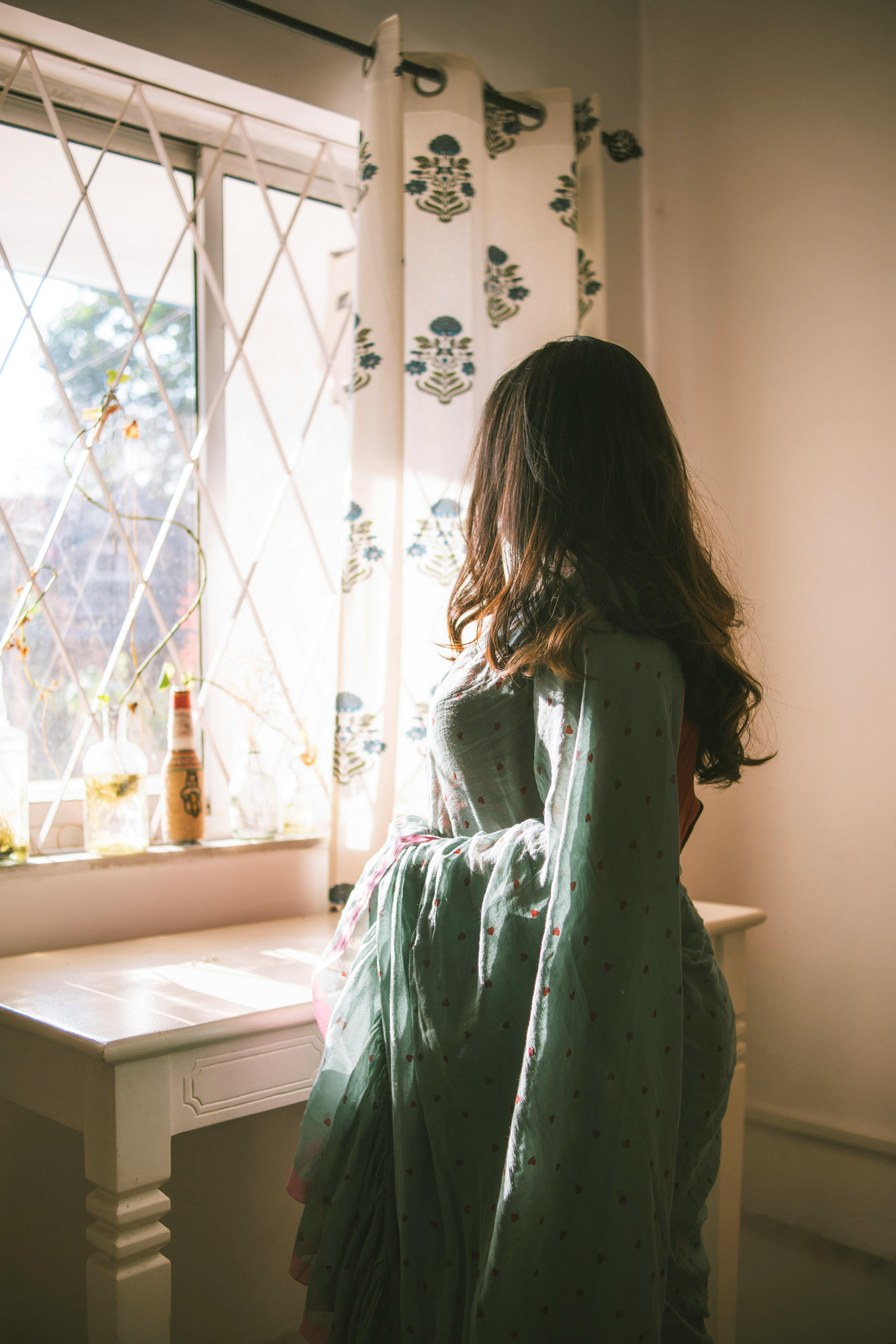 Gratis Una donna con un elegante sari è in piedi con grazia vicino a una finestra, da cui filtra una tenue luce del sole che crea un'atmosfera serena. Foto a disposizione
