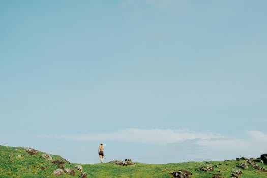 A single hiker explores a wide open green field under a clear blue sky.