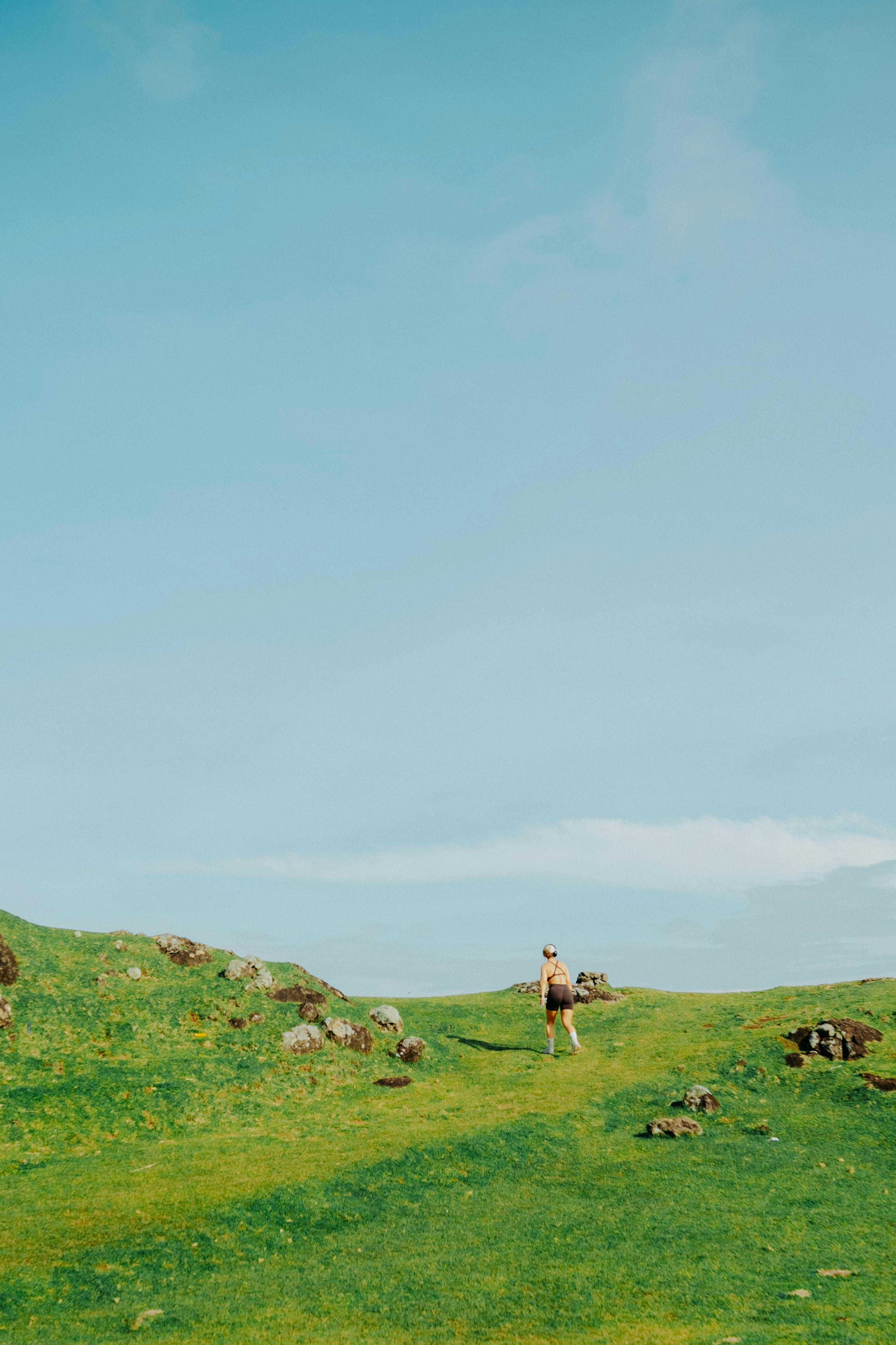 Ücretsiz Tek başına bir kadın, masmavi bir gökyüzünün altında yemyeşil bir yamaçta yürüyüş yapıyor. Stok Fotoğraflar