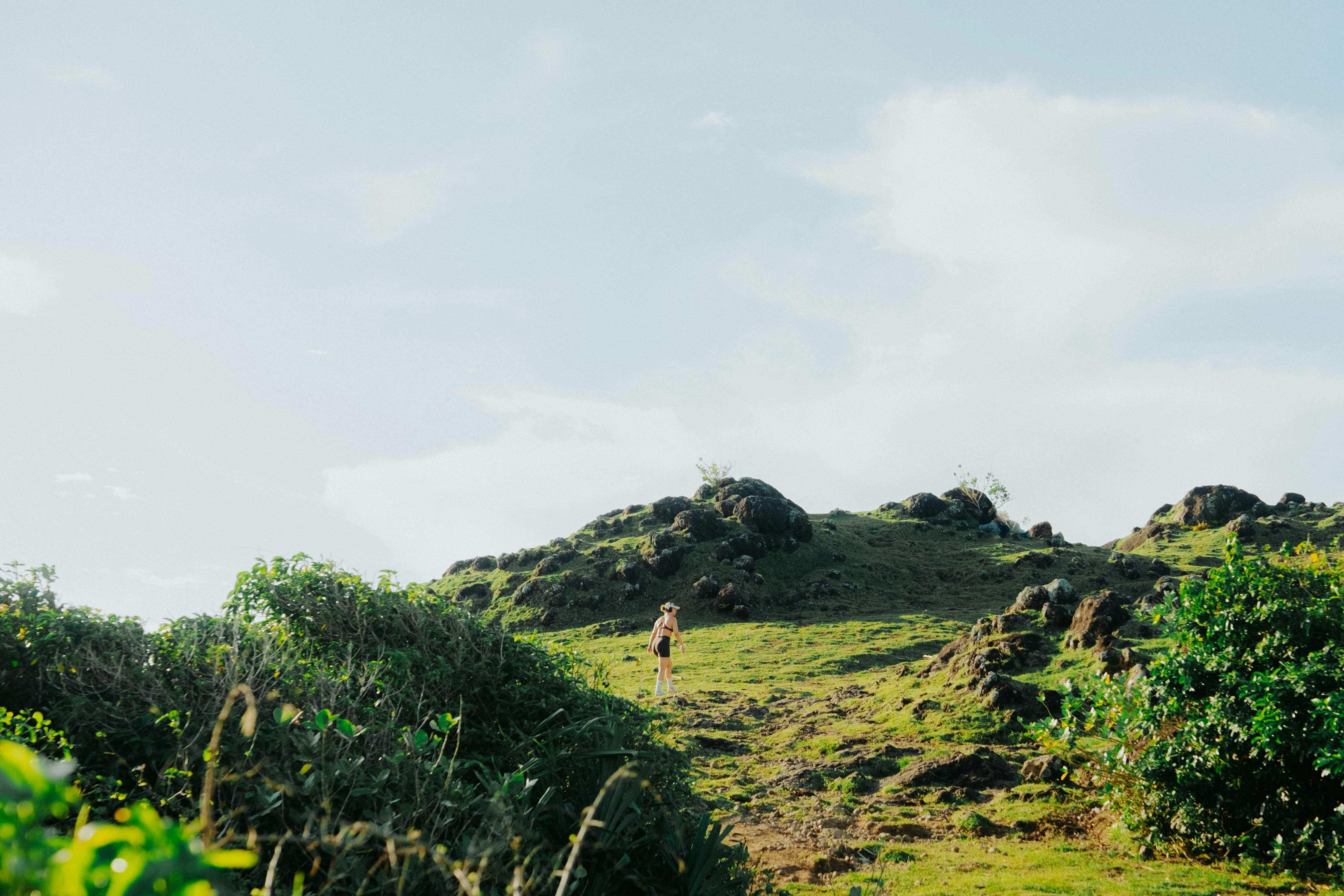 Gratis Una persona sale su una collina erbosa circondata da un terreno roccioso sotto un cielo azzurro brillante. Foto a disposizione