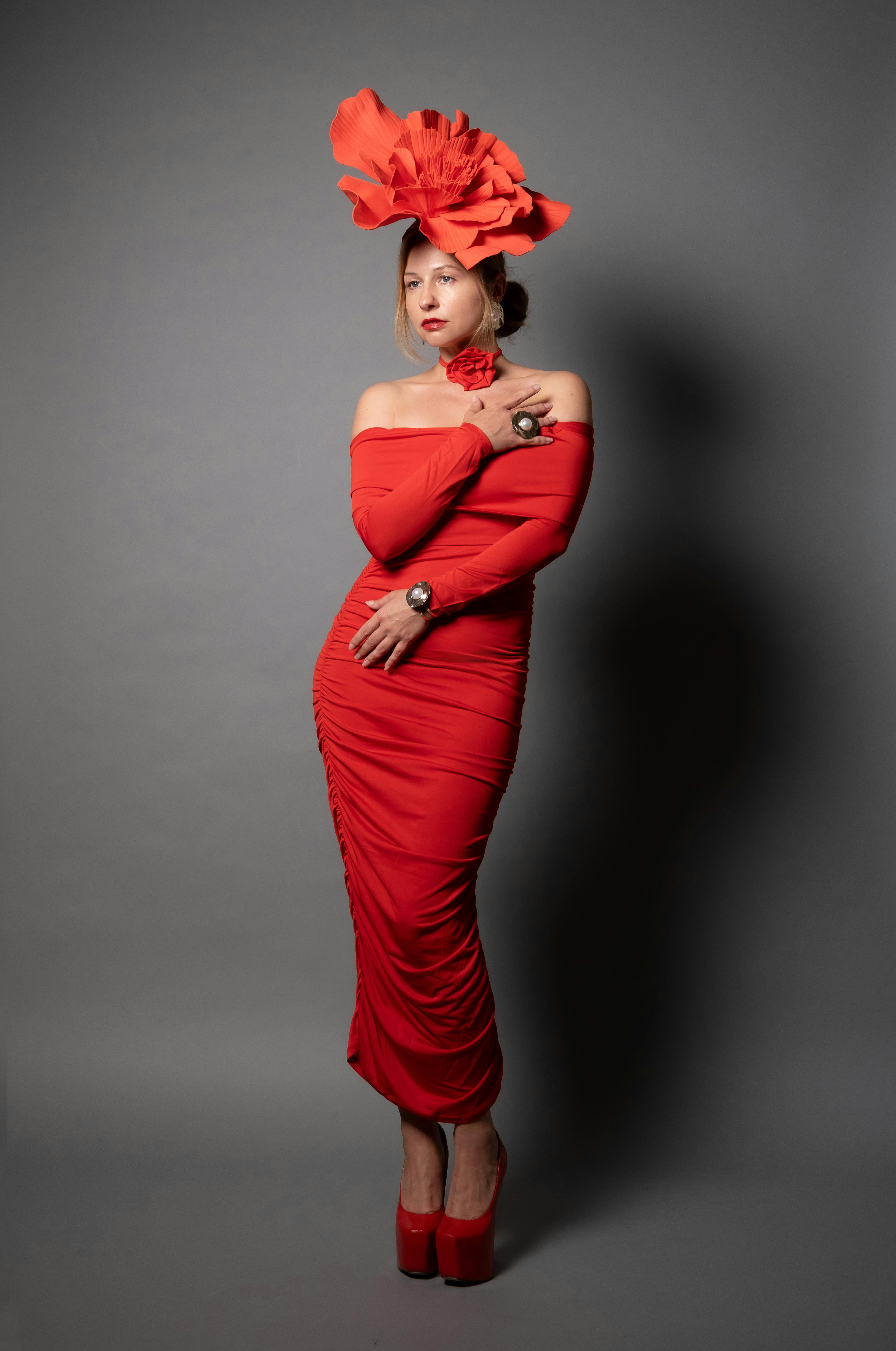 ฟรี หญิงสาวผู้มีสไตล์ในชุดเดรสสีแดงสดใส พร้อมหมวกประดับศีรษะที่สะดุดตา ยืนอยู่บนฉากหลังสีเทา คลังภาพถ่าย