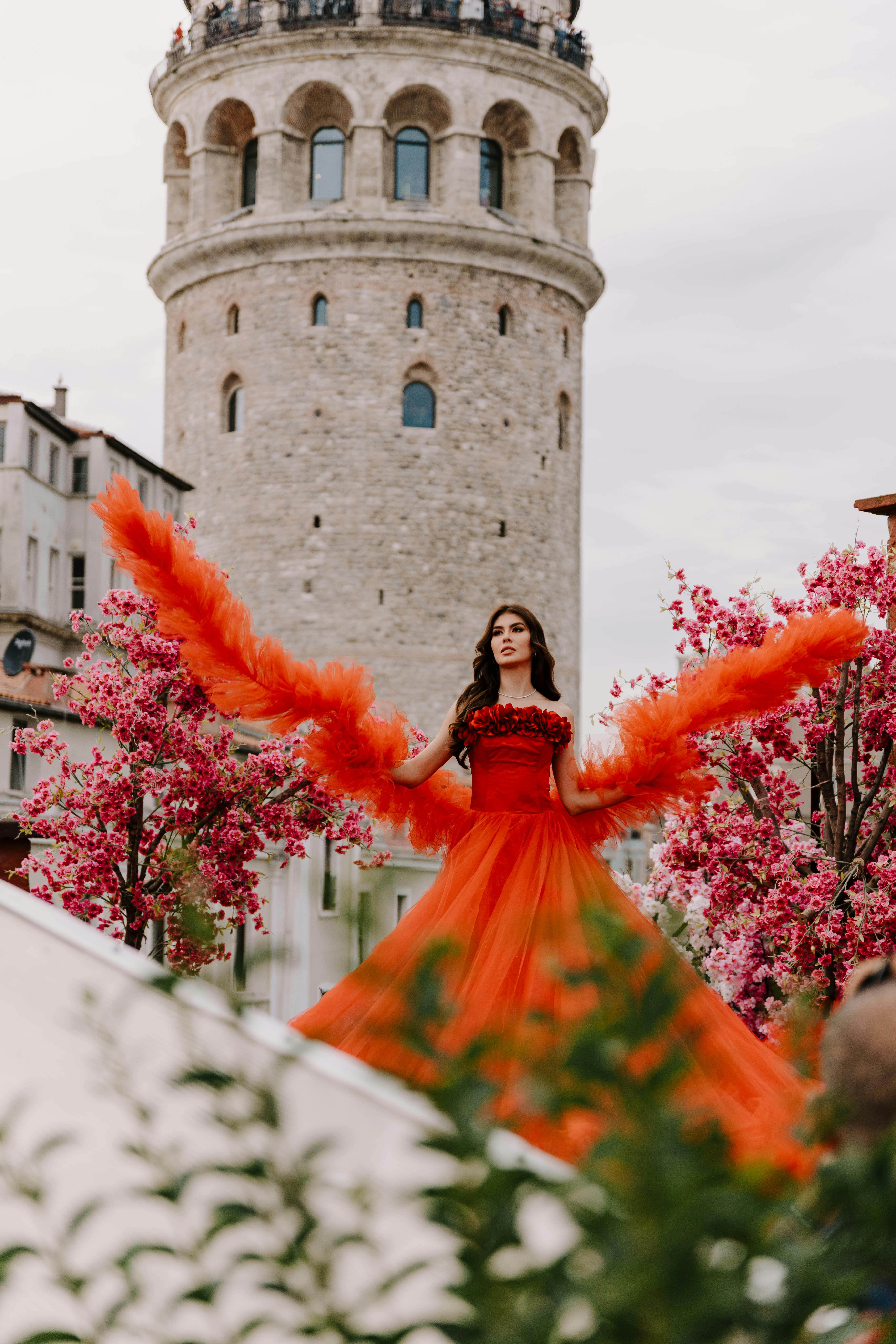 Gratis Una donna elegante con un abito rosso fluente posa con grazia sullo sfondo della Torre di Galata, circondata da fiori di ciliegio. Foto a disposizione