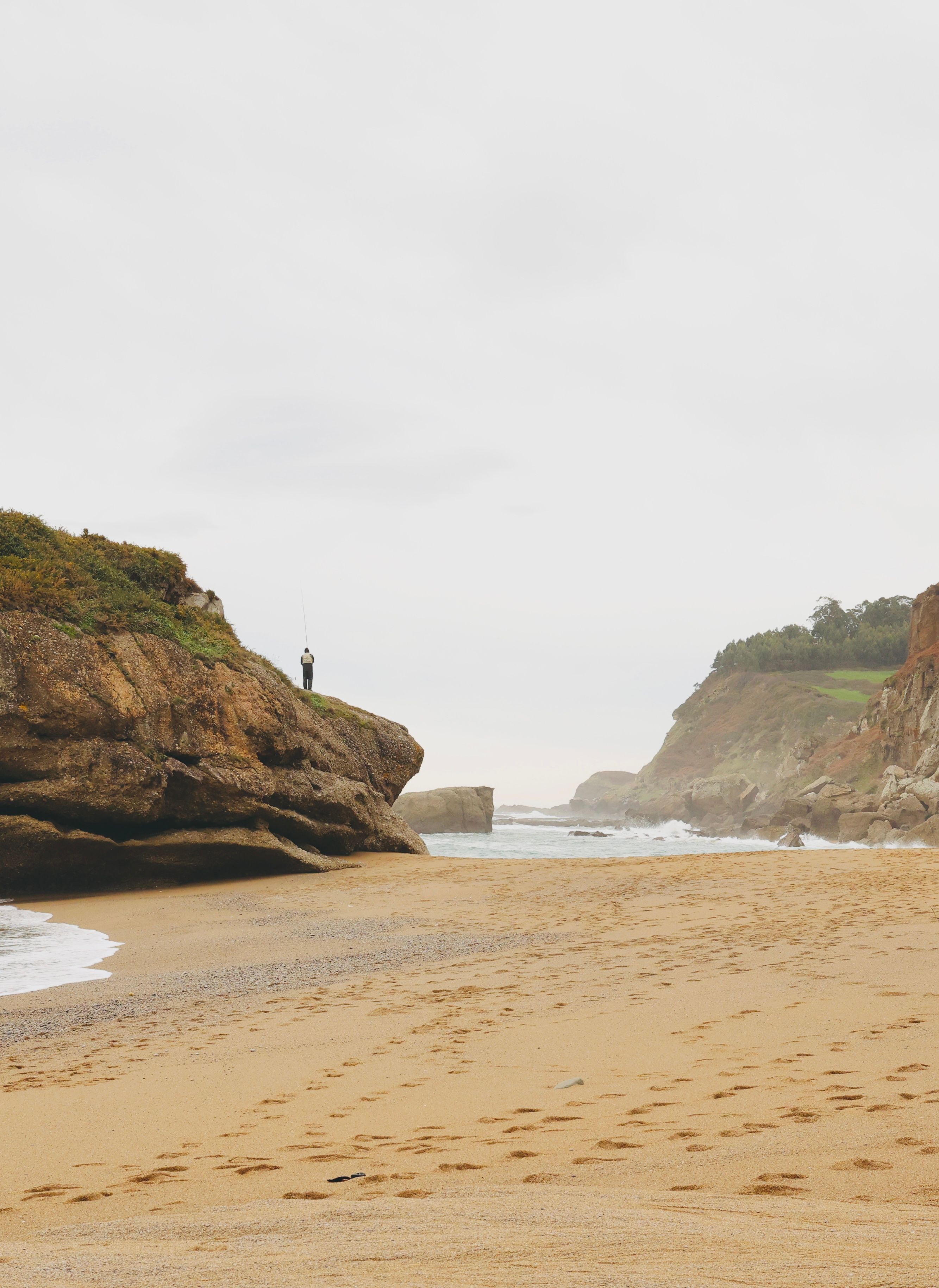 A lone fisherman stands atop a rocky outcrop on a serene, sandy beach, facing the ocean waves.
