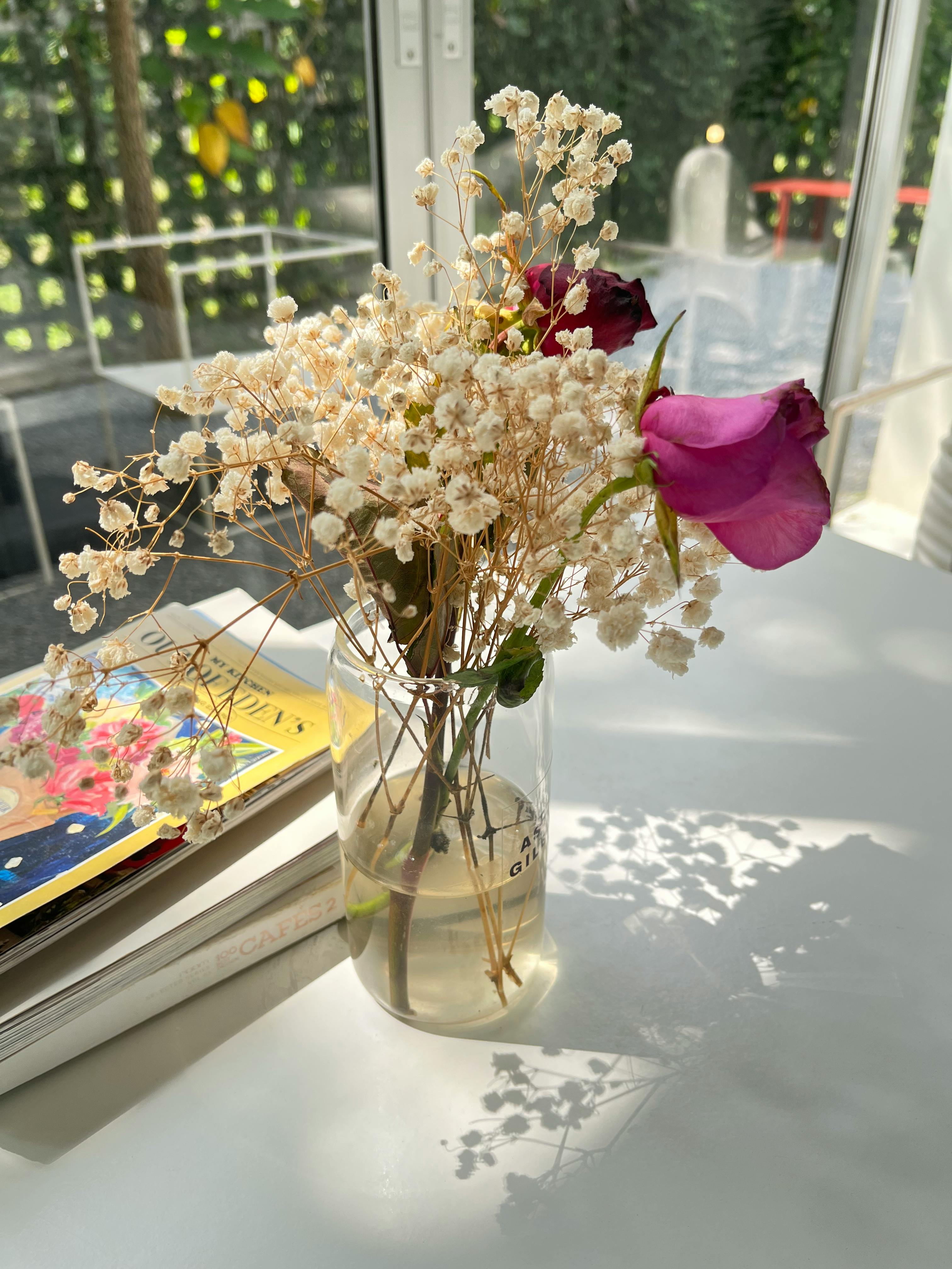 Ilmainen Kirkas kukka-asetelma lasimaljakossa auringonvalolla valaistulla pöydällä sisätiloissa näkyvillä kirjan kansilla. Kuvapankkikuva