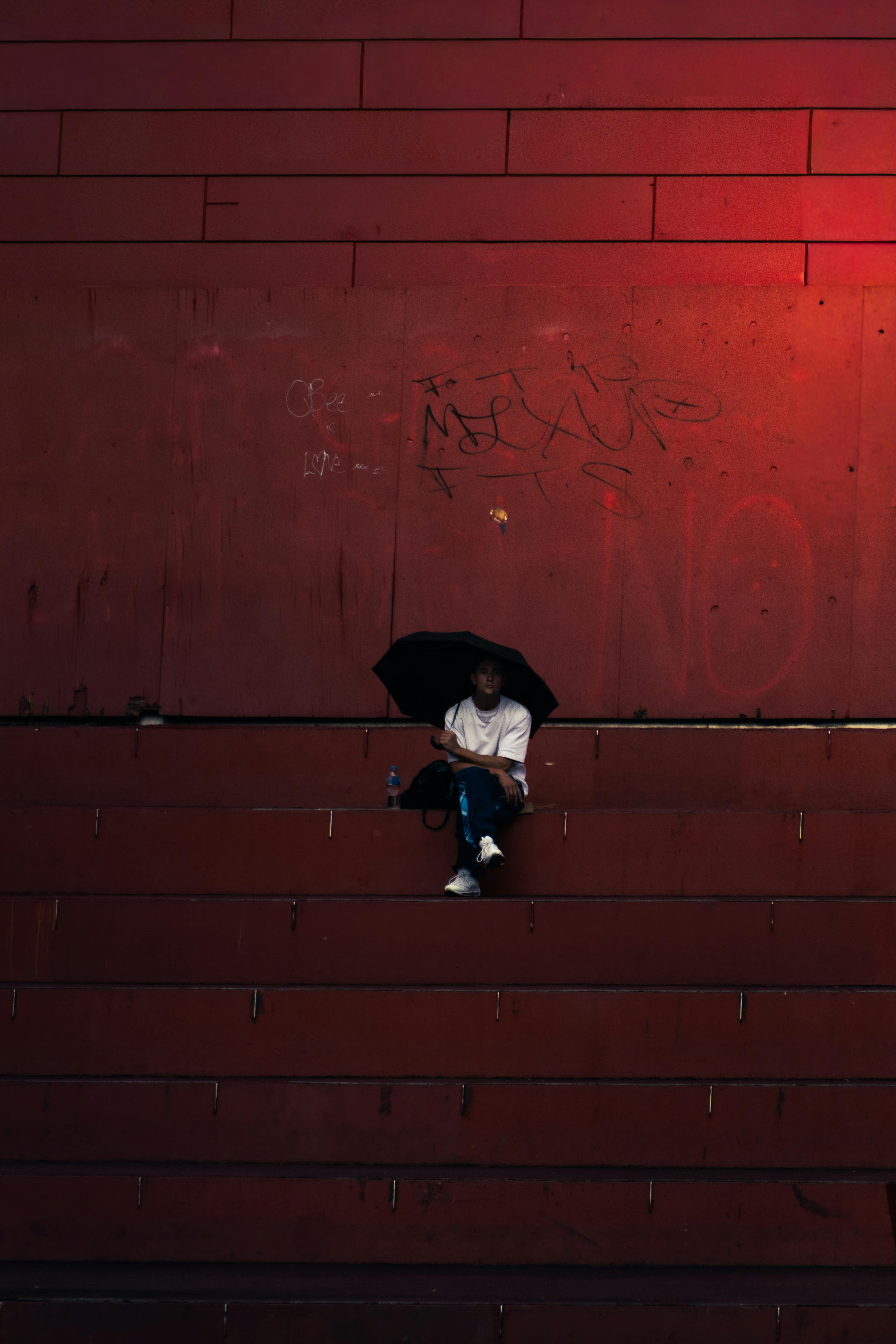 бесплатная Человек сидит в одиночестве на красных ступеньках, держа в руках зонтик, создавая впечатляющую городскую сцену. Стоковое фото