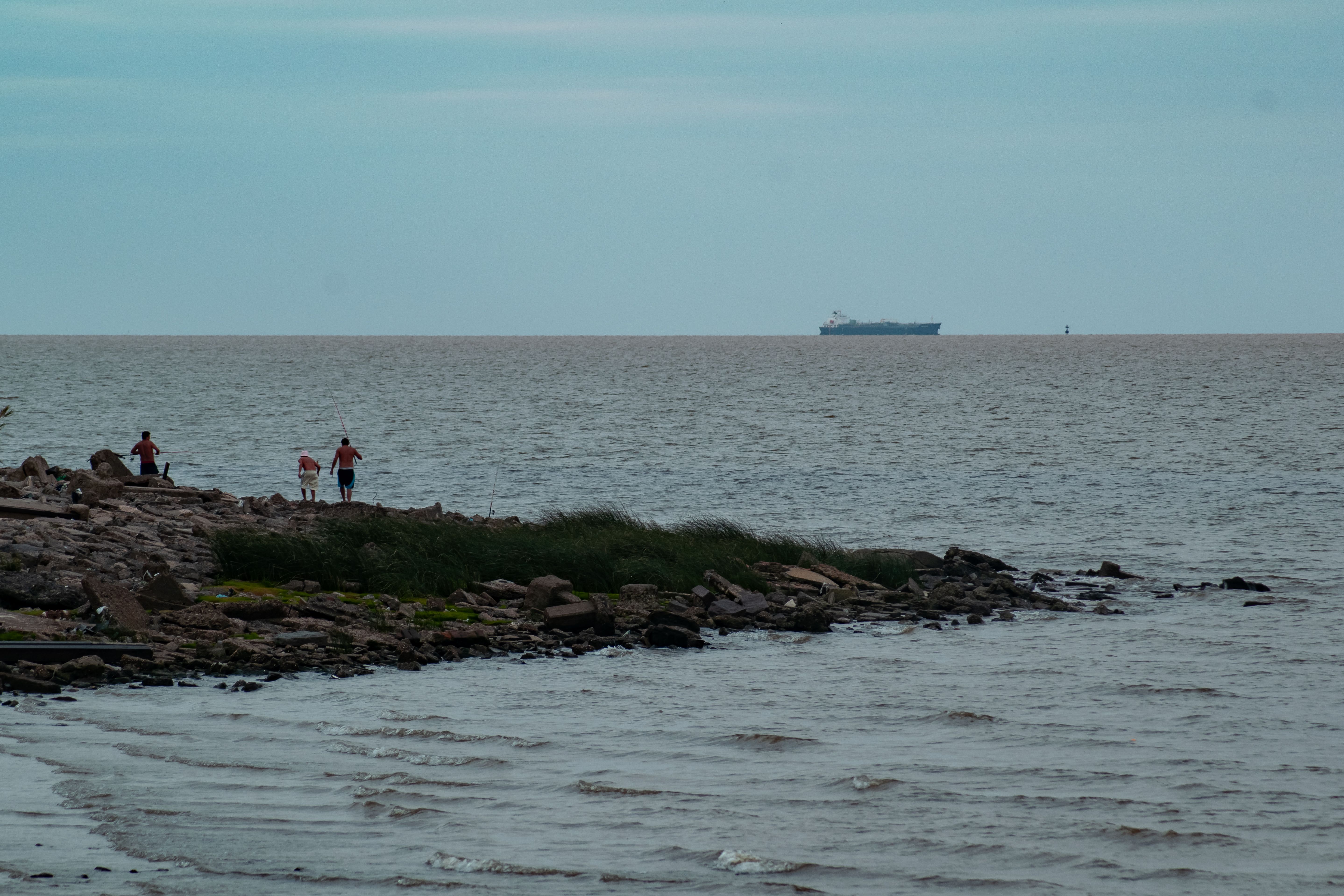Serene coastal scene with fishing and a distant cargo ship under an open sky.