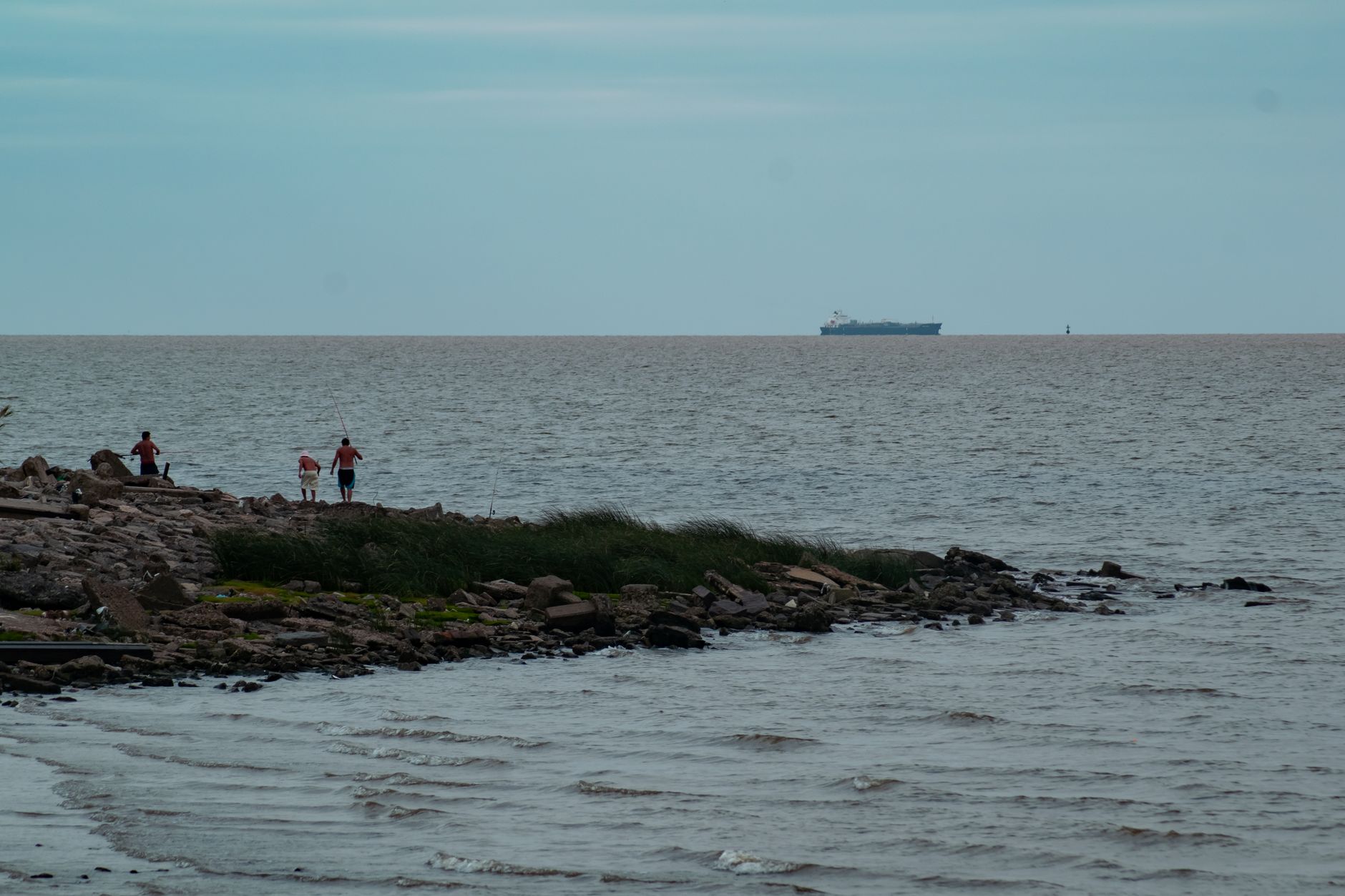 Serene coastal scene with fishing and a distant cargo ship under an open sky.