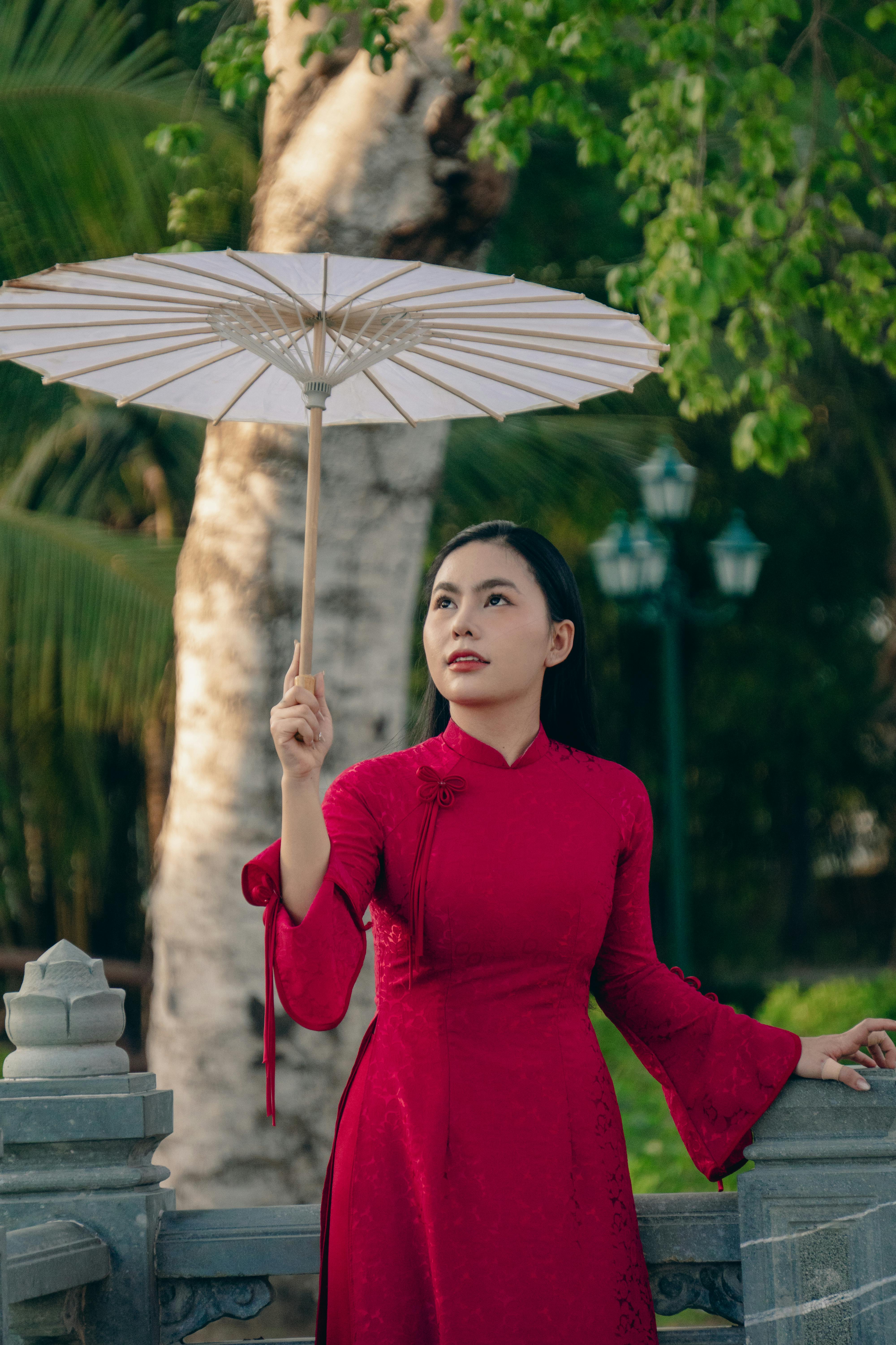 Elegant Vietnamese woman in a red Ao Dai holding a white umbrella in a serene outdoor setting.