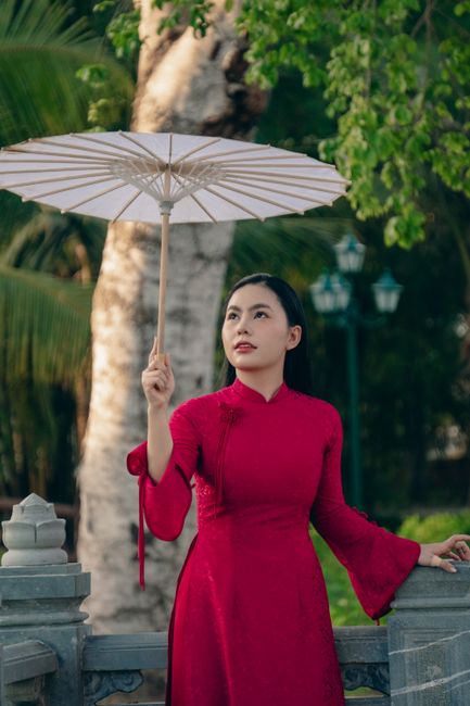 Elegant Vietnamese woman in a red Ao Dai holding a white umbrella in a serene outdoor setting.