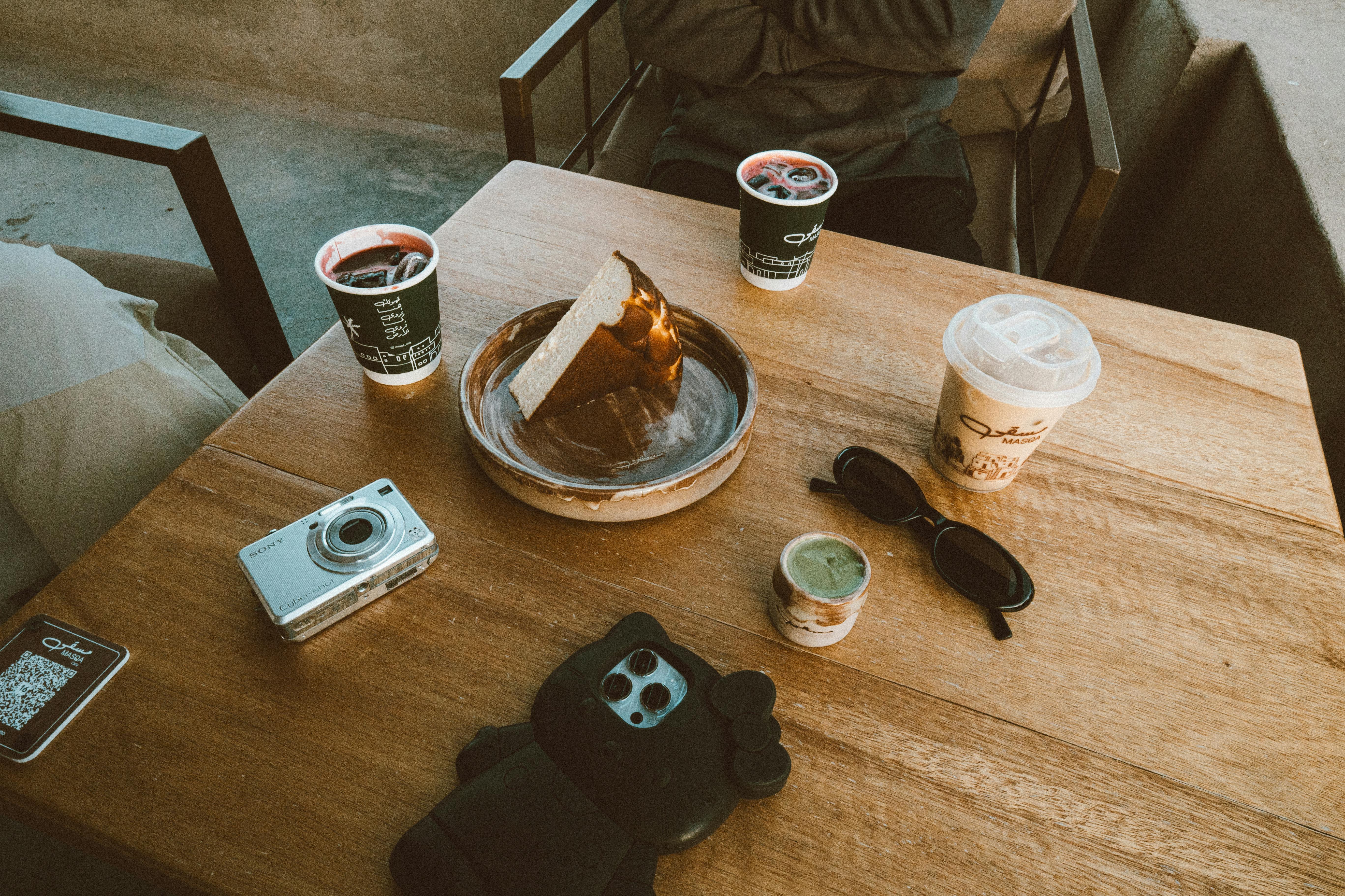 Free A relaxing scene with coffee, cake, and a camera on a wooden table at an Oman cafe. Stock Photo